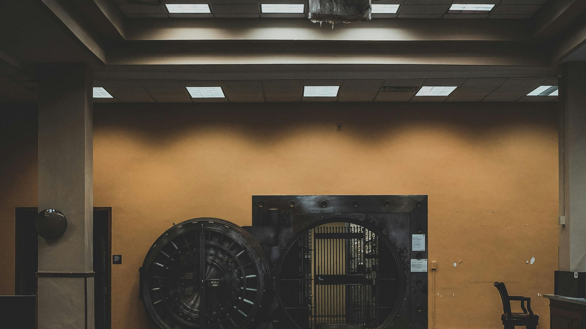 Old bank vault door in a dimly lit room, with an orange wall and rows of ceiling lights.