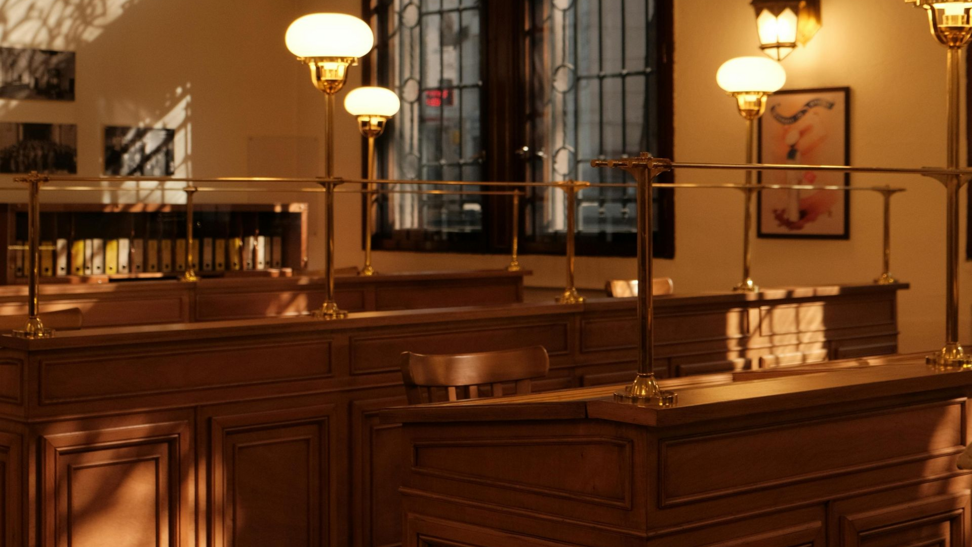 Wooden bank teller booths with brass railings and lamps, in a room with a window.