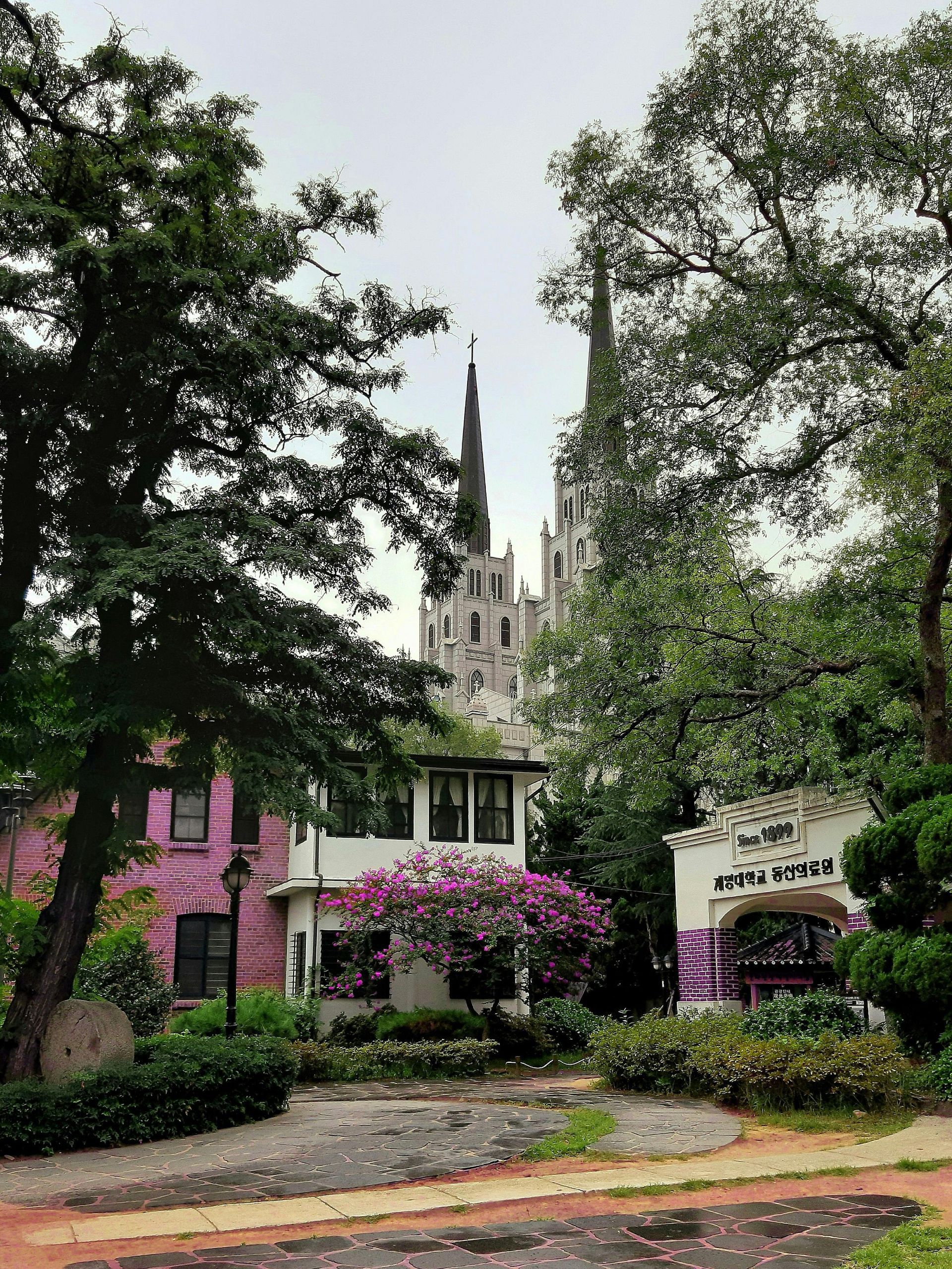 Colorful buildings and trees in front of a tall, gothic-style structure under a cloudy sky.