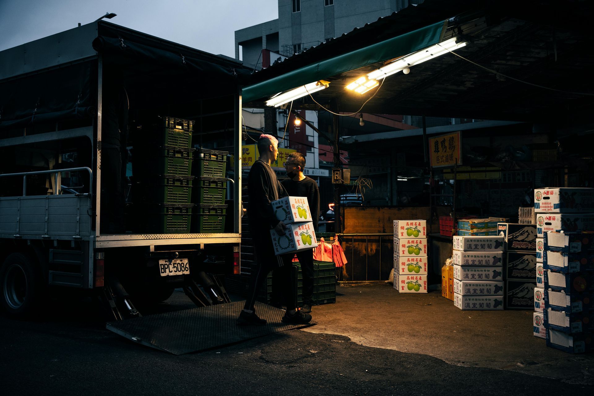 Truck unloading produce at a market stall; boxes stacked nearby. Two figures by the truck. Evening lighting.