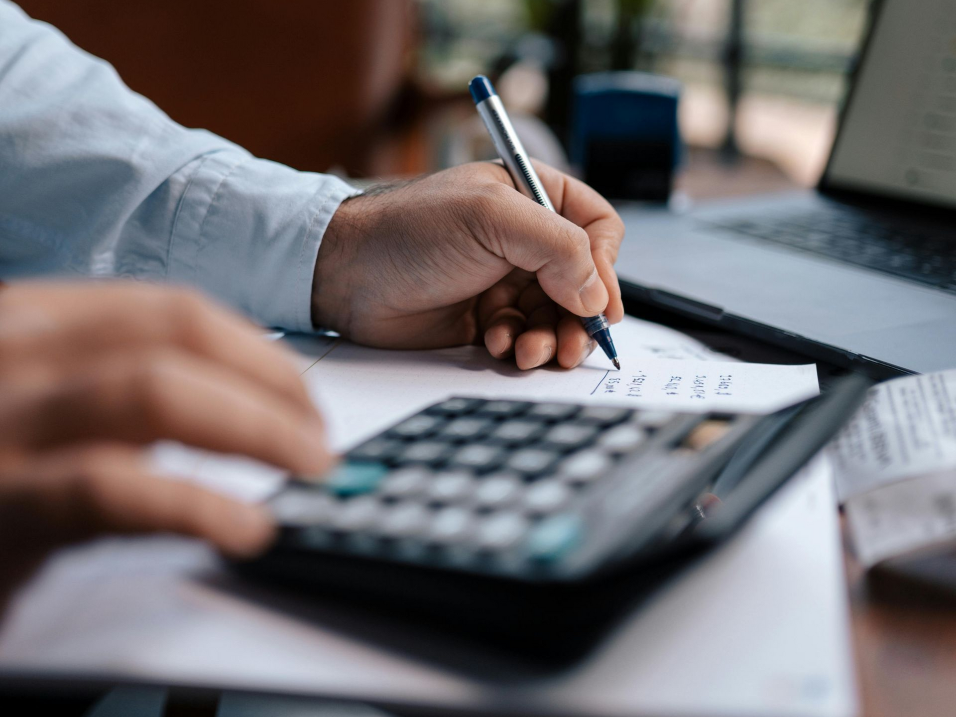 Person writing with pen, calculating with calculator, near a laptop and papers.