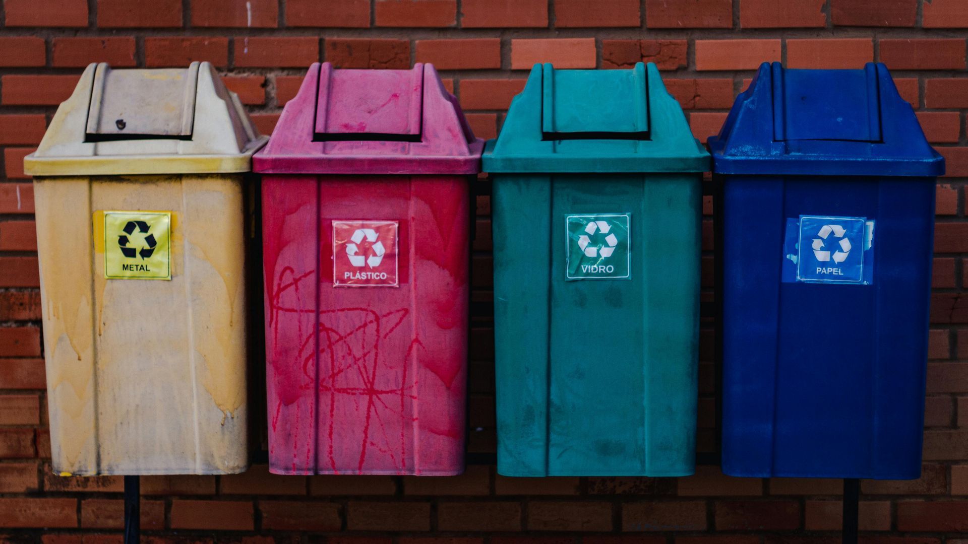 Four colorful recycling bins against a brick wall: yellow, pink, teal, and blue. Each has a recycling symbol.