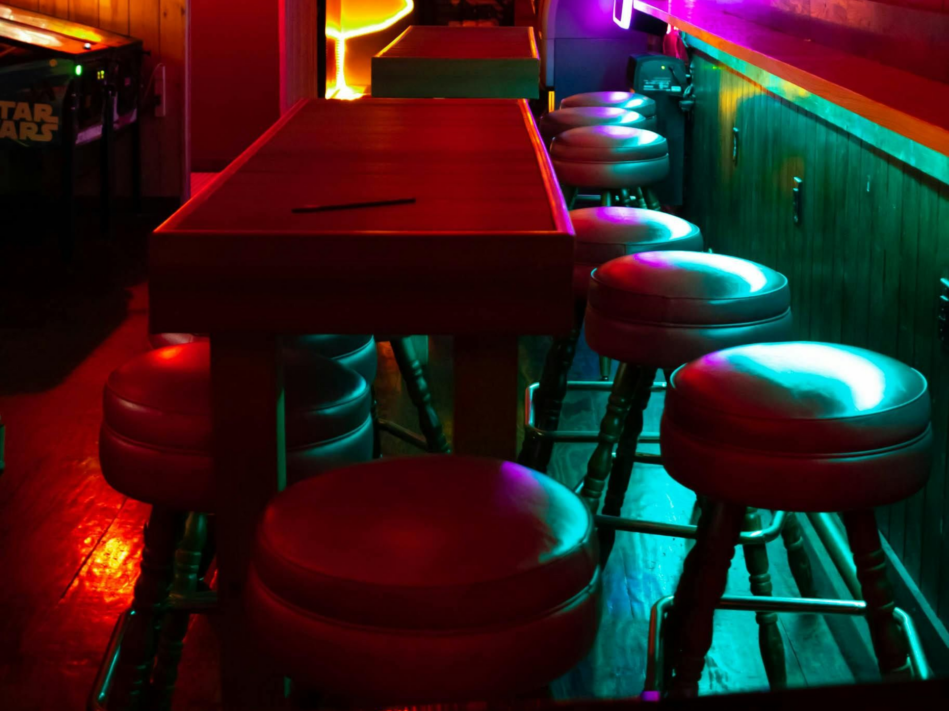 Bar with red-lit wooden table, stools, and green-lit wall; dimly lit setting.