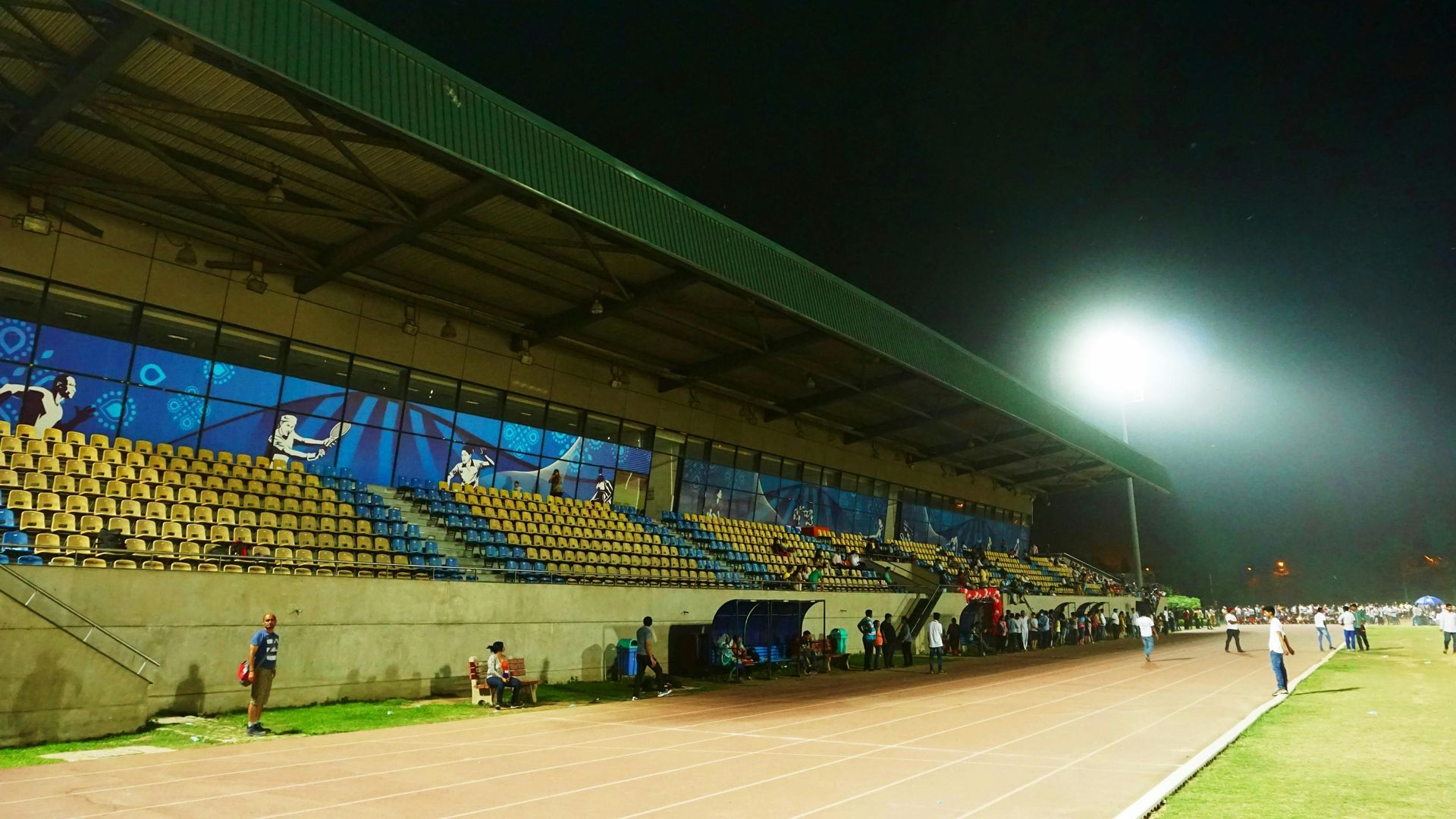 Nighttime view of a stadium with running track, stands, and lighting. People gather.