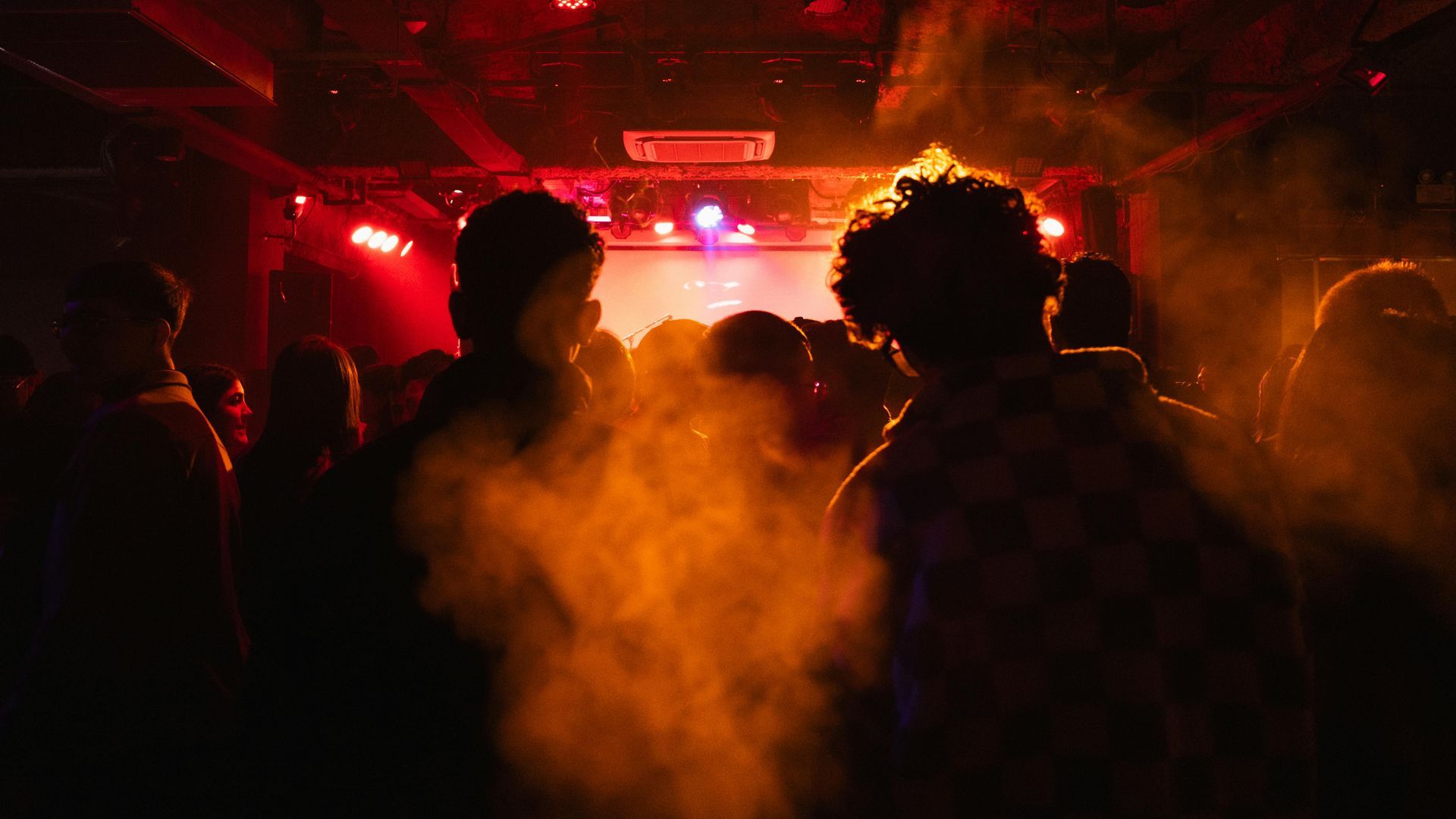 People in a smoky, red-lit nightclub; silhouettes of dancers, stage lights in background.