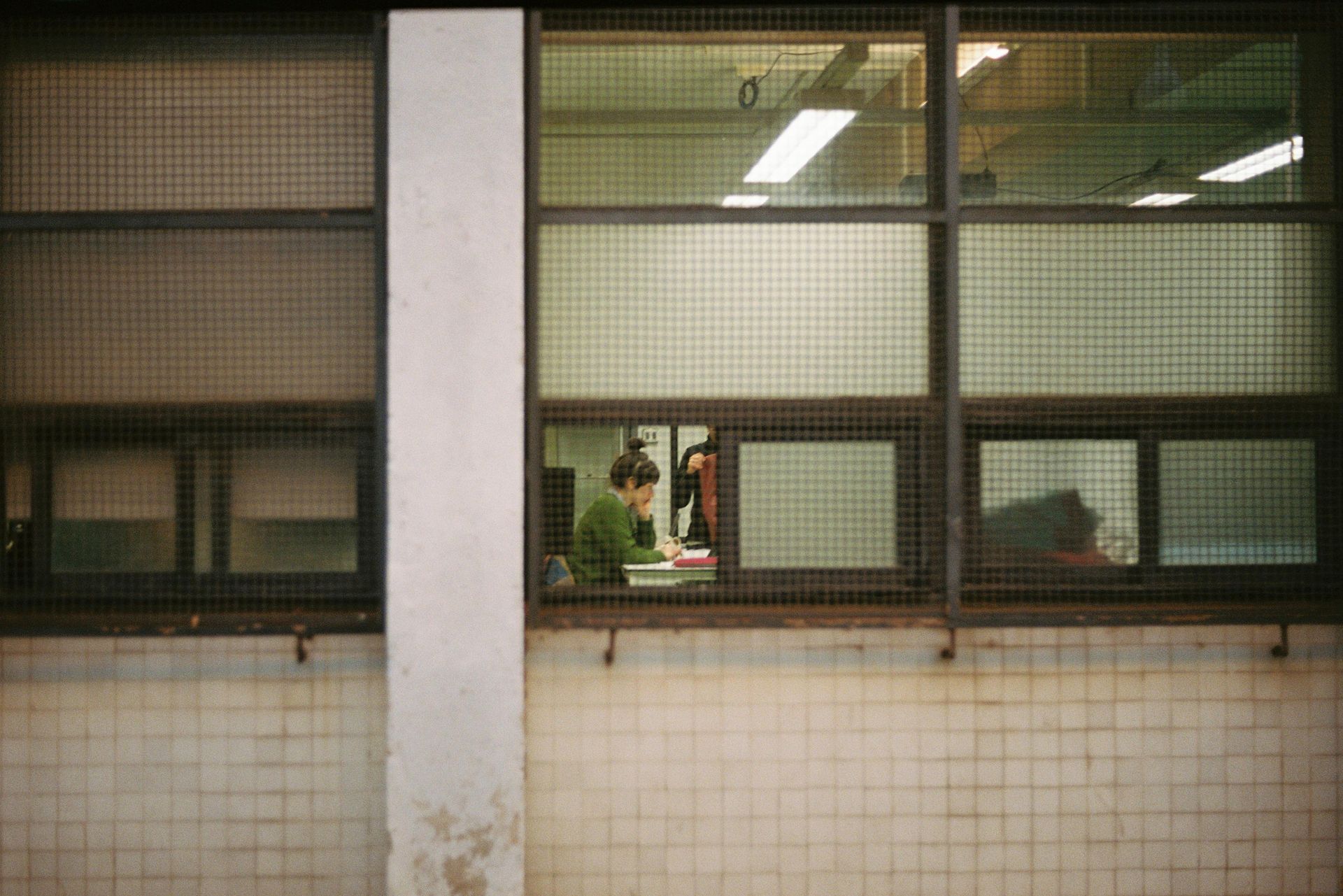 A person in a green sweater sits studying at a desk inside a windowed office building with a tiled exterior wall.