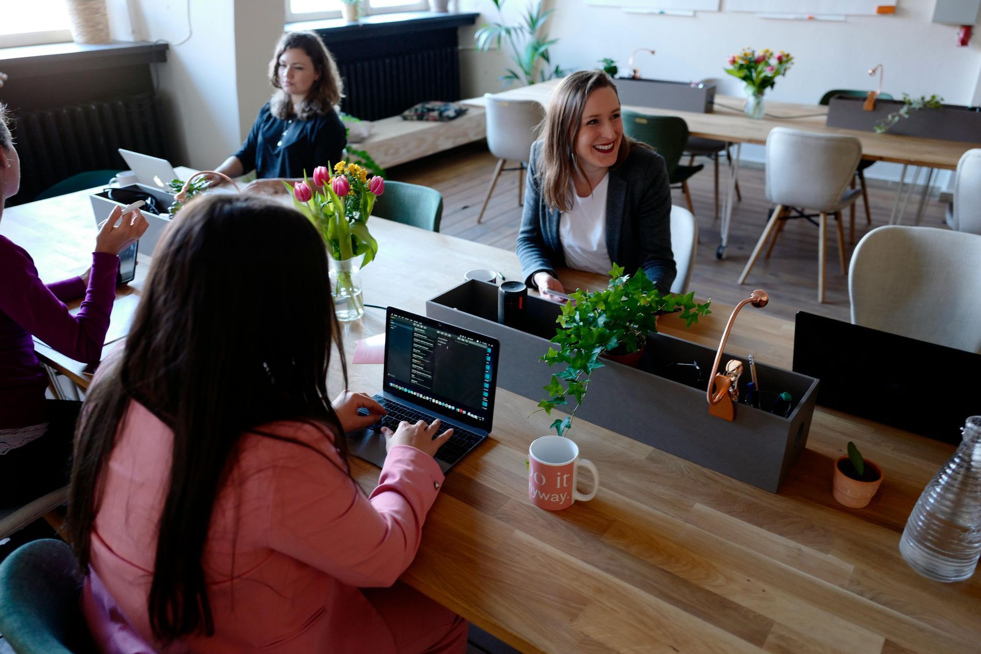 People in an office at a table. Woman types on laptop; another woman smiles. Flowers and plants on the table.