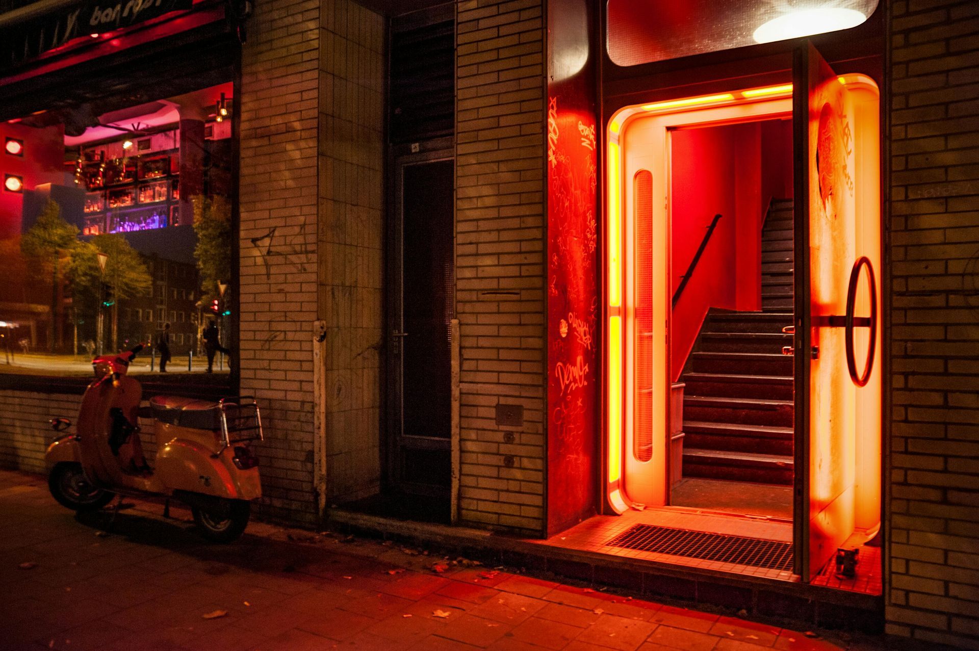 Night scene: red-lit nightclub entrance, brick building, scooter parked outside. Stairway leads upwards.