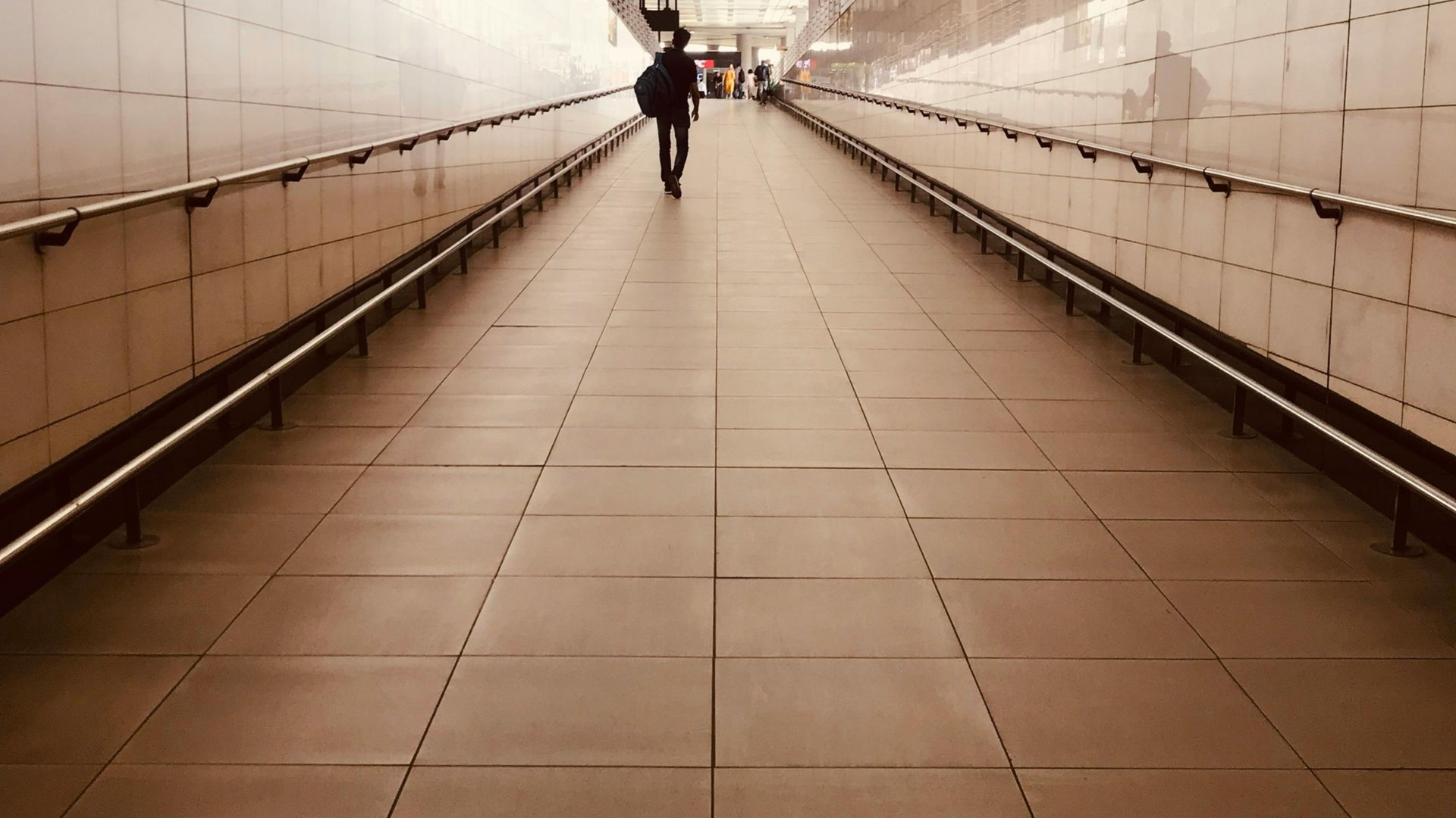 Person walking down a long, tiled hallway with handrails; muted brown tones.