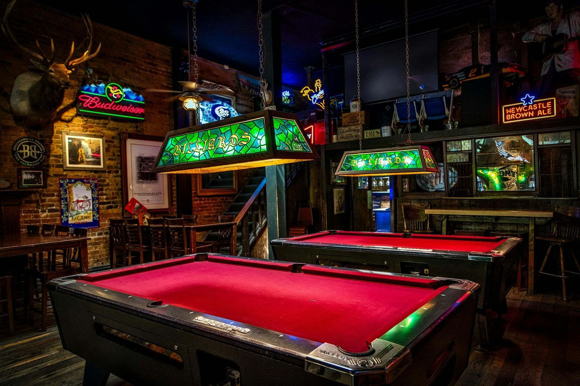 Two red-felted pool tables in a dimly lit bar, with hanging lights, exposed brick, and a deer head on the wall.