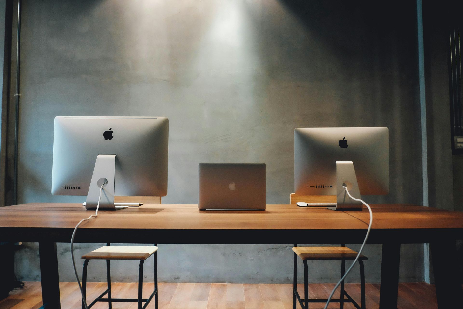 Two Apple monitors and a laptop on a wooden desk with two stools in front of a concrete wall.