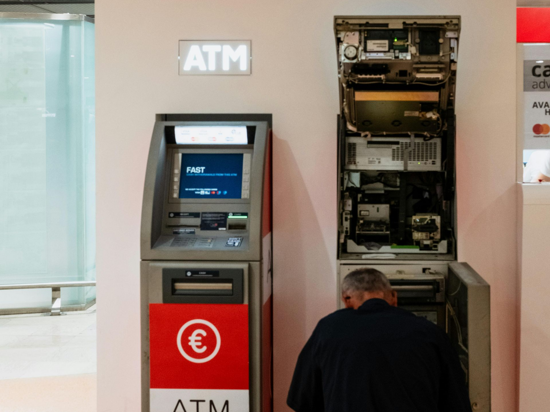 Man working on an open ATM. Another ATM next to it. Indoor, white walls with red accents.