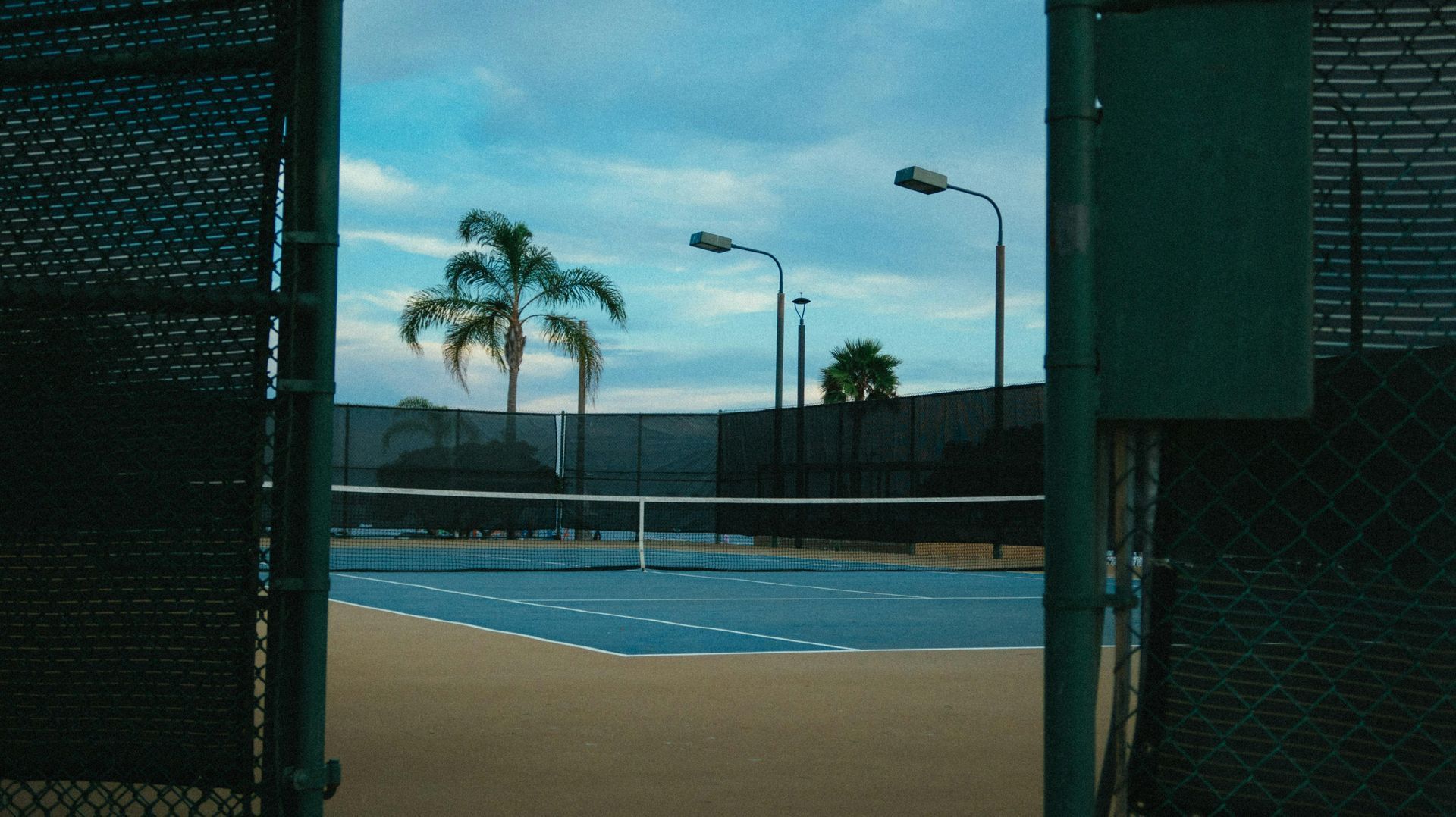 Tennis court viewed through an open gate, blue surface, palm trees, overcast sky.