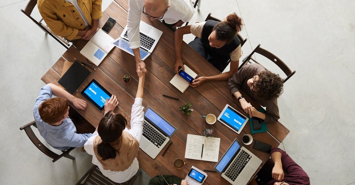 People shaking hands at a wooden table with laptops, tablets, and coffee cups.