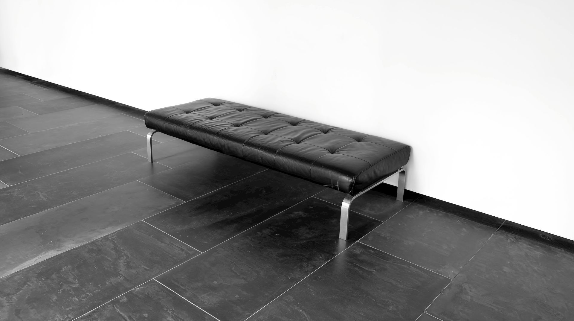 Black bench with tufted seat, metal legs, against a white wall and dark tiled floor.