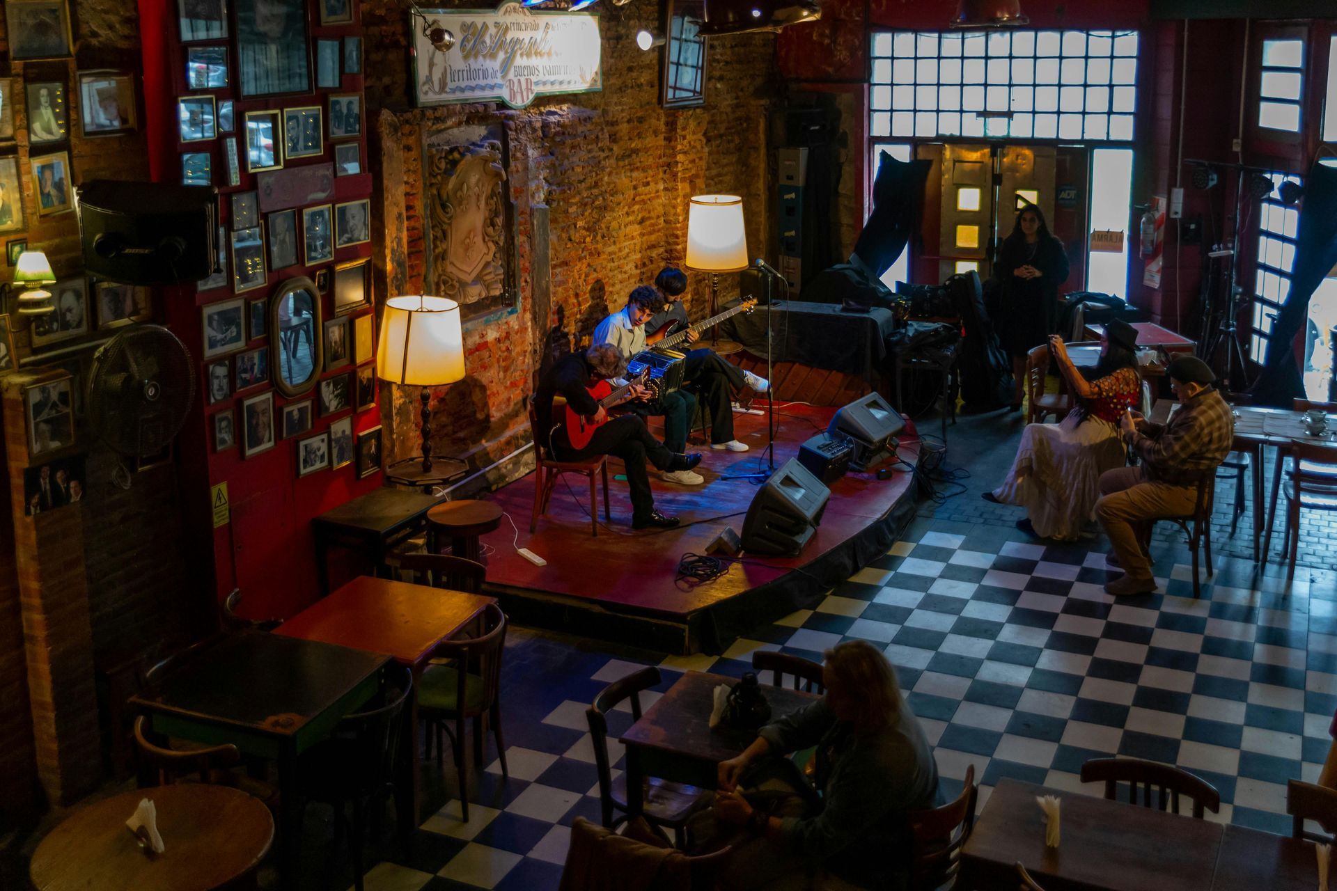 A band performs on a small stage in a dimly lit bar with checkered floors and framed photos on the walls.