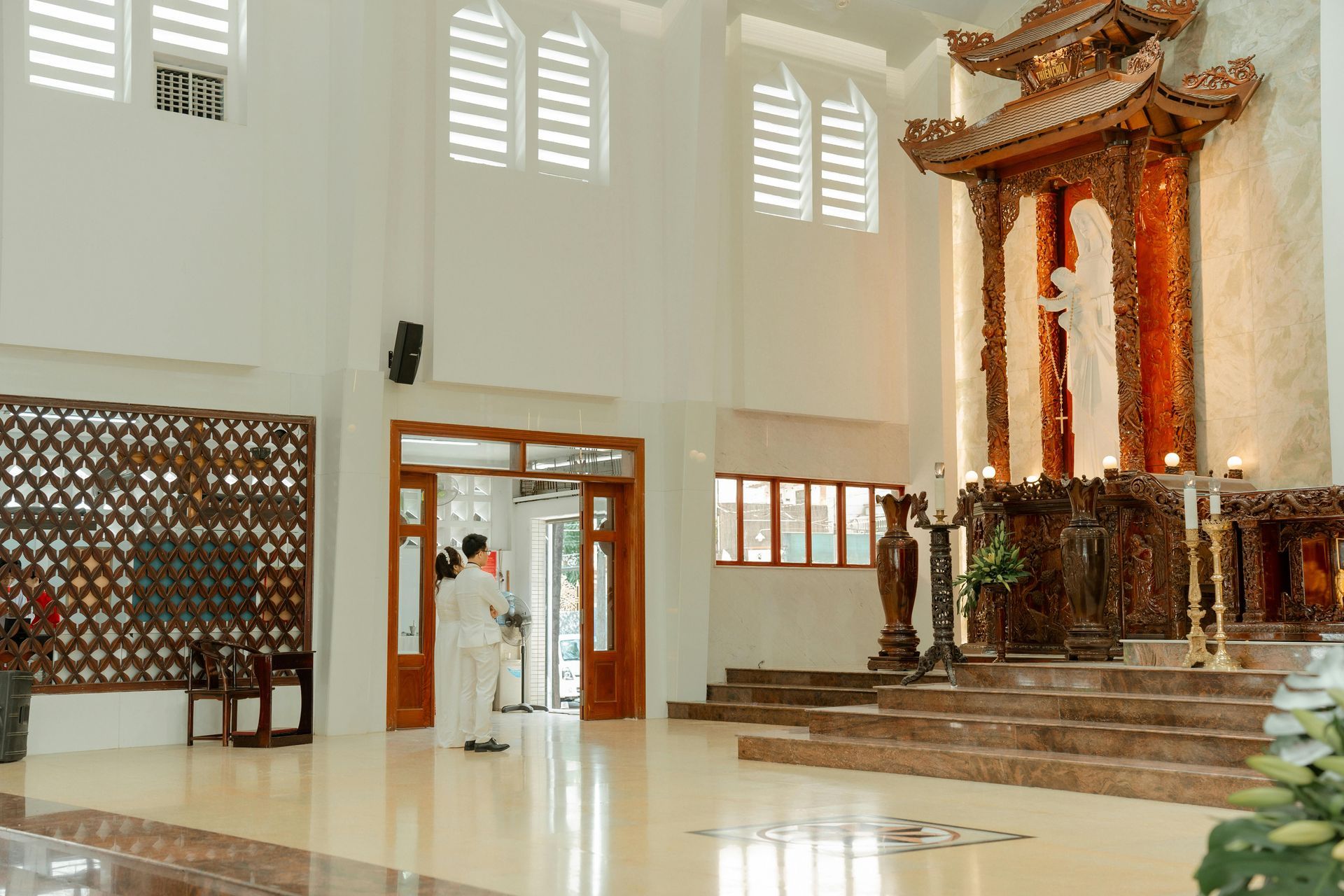 Interior view of a church with an ornate altar, a person stands at the open doorway, and a tiled floor.