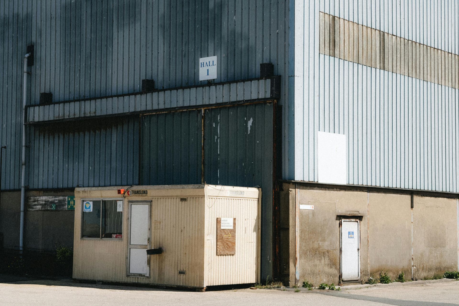 A small wooden security booth next to a large industrial building with a white door and a sign.