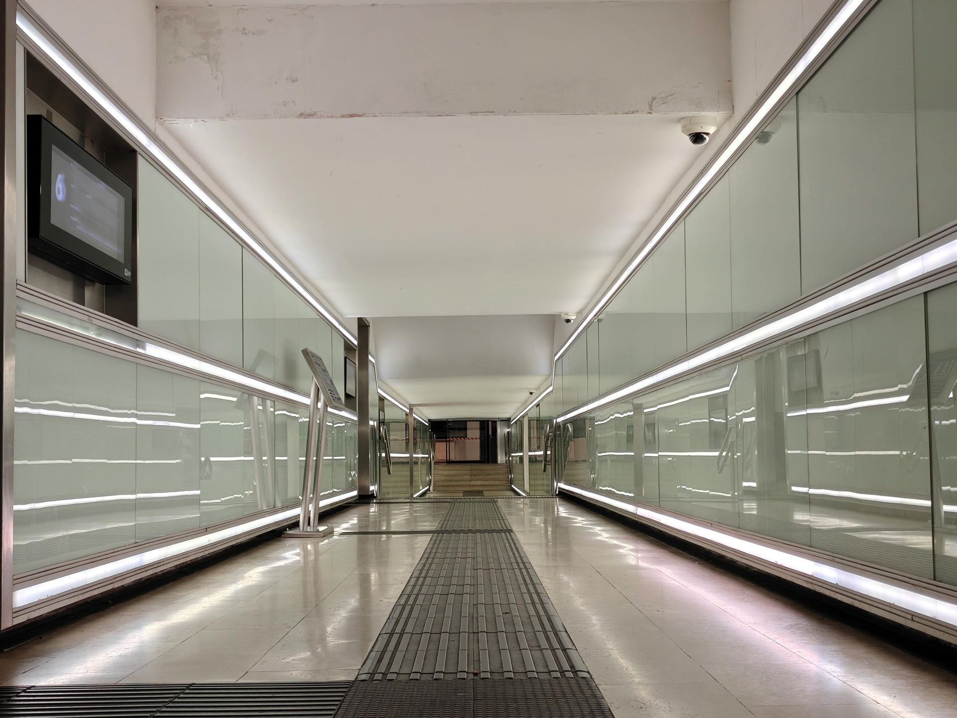 Long, bright hallway with glass walls, overhead lighting, and metal floor panels.
