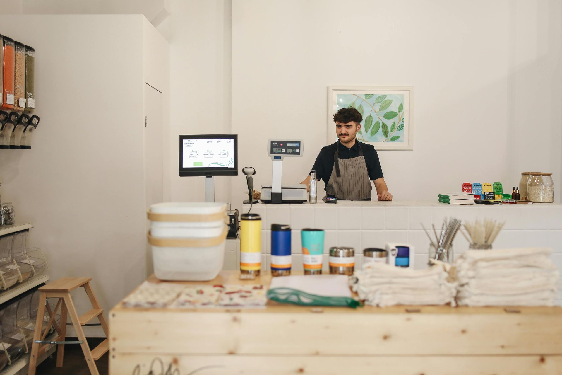 Man behind counter in eco-friendly shop, smiling, with scale, reusable containers, and merchandise displayed.