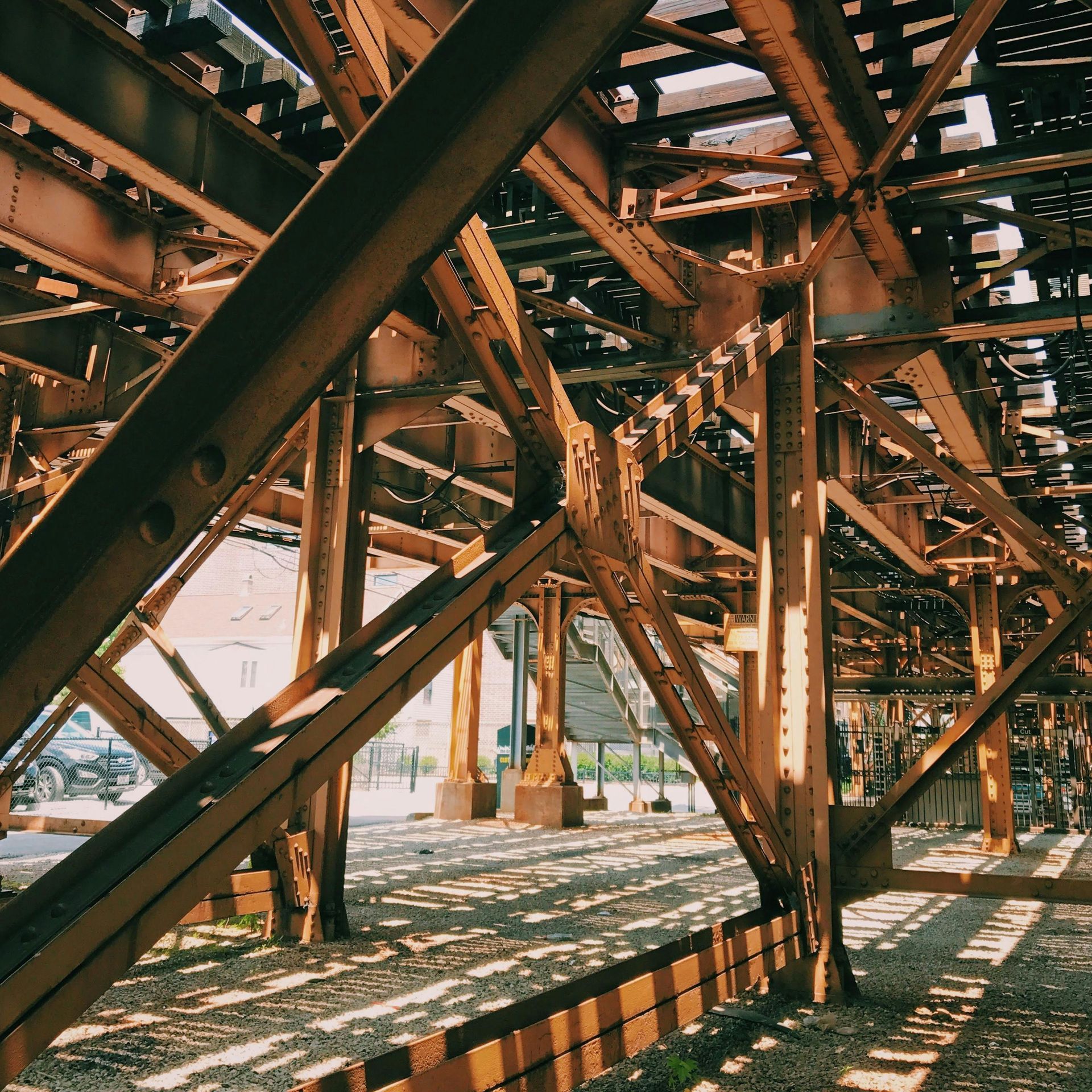 Rust-colored metal beams and supports of an elevated train structure casting shadows on the ground below.