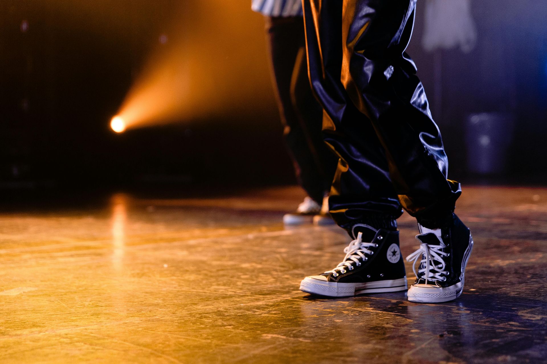 Person in black pants and high-top Converse sneakers dancing on stage, spotlight illuminating them.