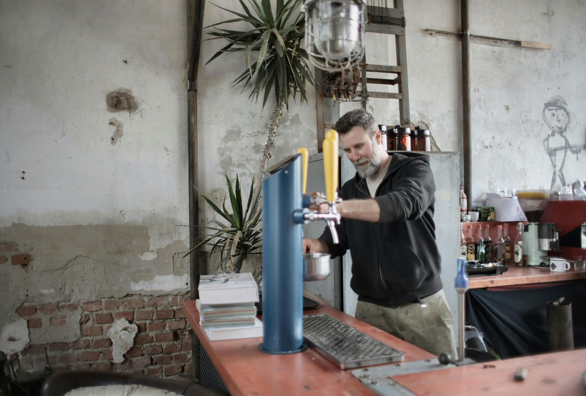 Man pouring beer from a blue tap at a bar. Weathered walls, potted plants, and simple decor.