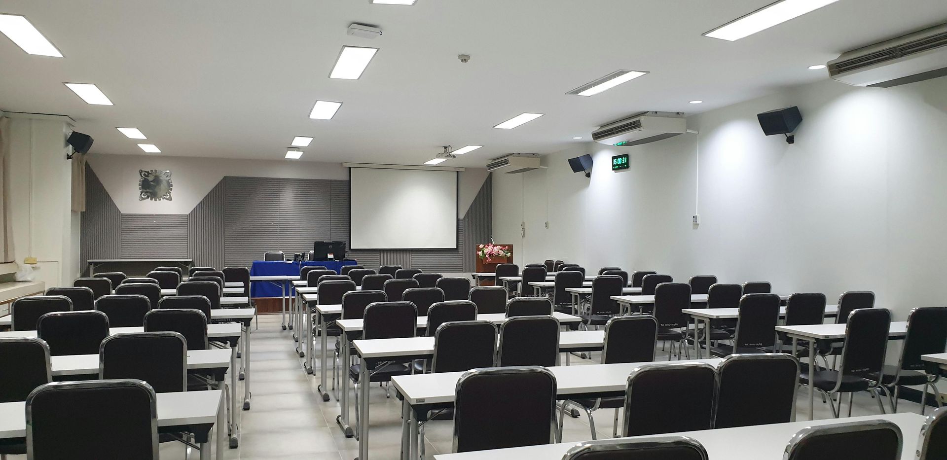 A brightly lit, empty classroom with rows of desks and black chairs facing a projector screen at the front.