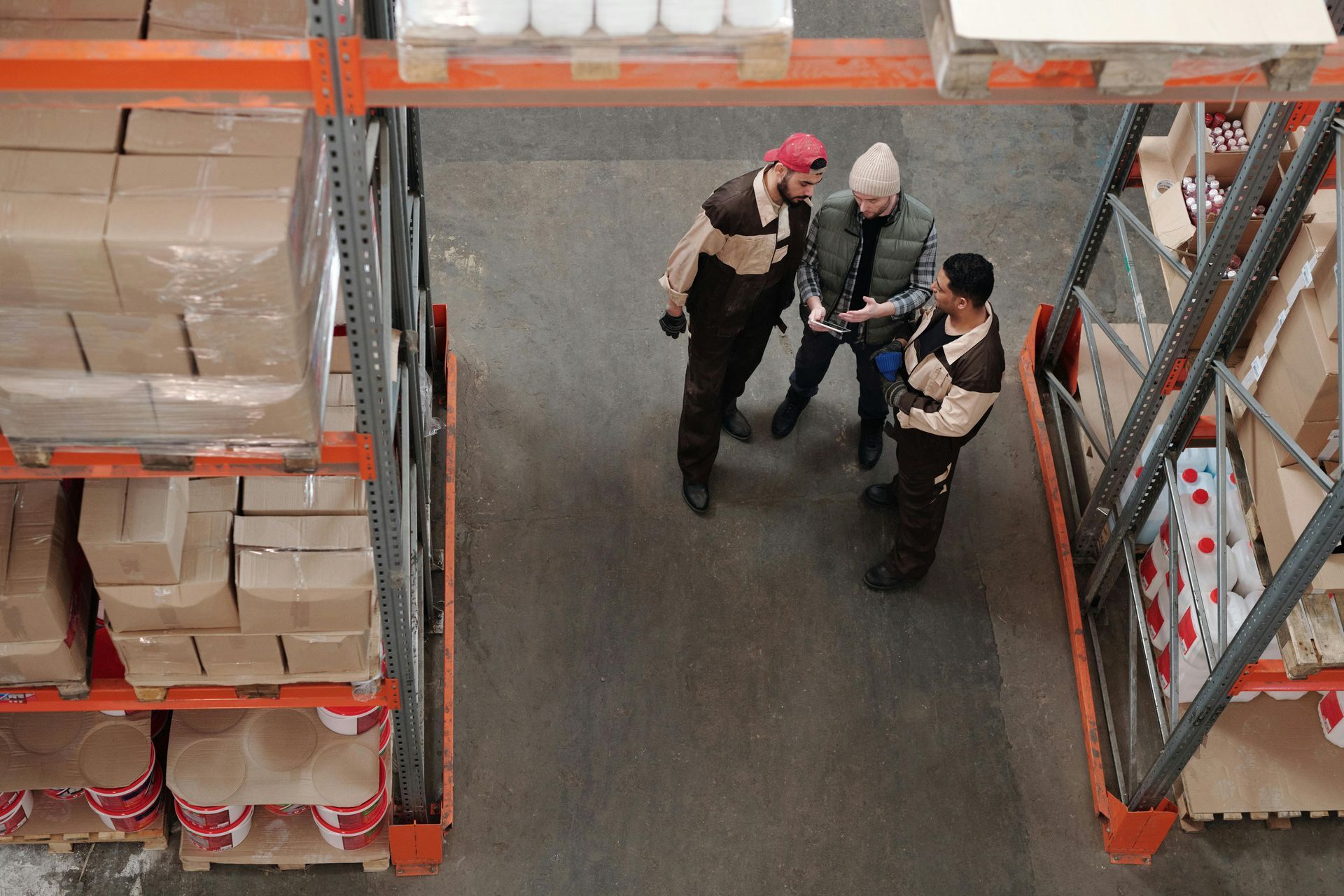 Three people in work uniforms consult a tablet in a warehouse setting, surrounded by shelves of packaged goods.