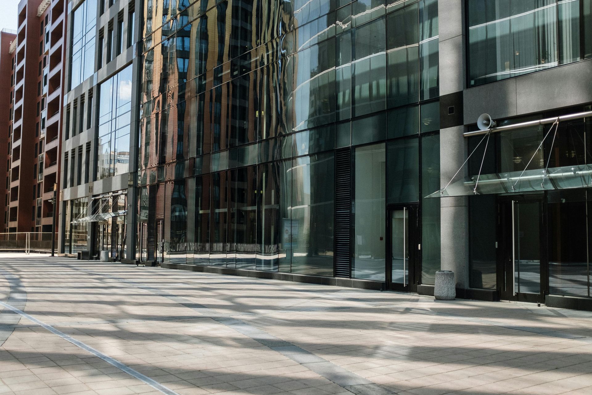 Modern glass-fronted office building and adjacent plaza with a distinctive grid-patterned shadow cast on the ground.