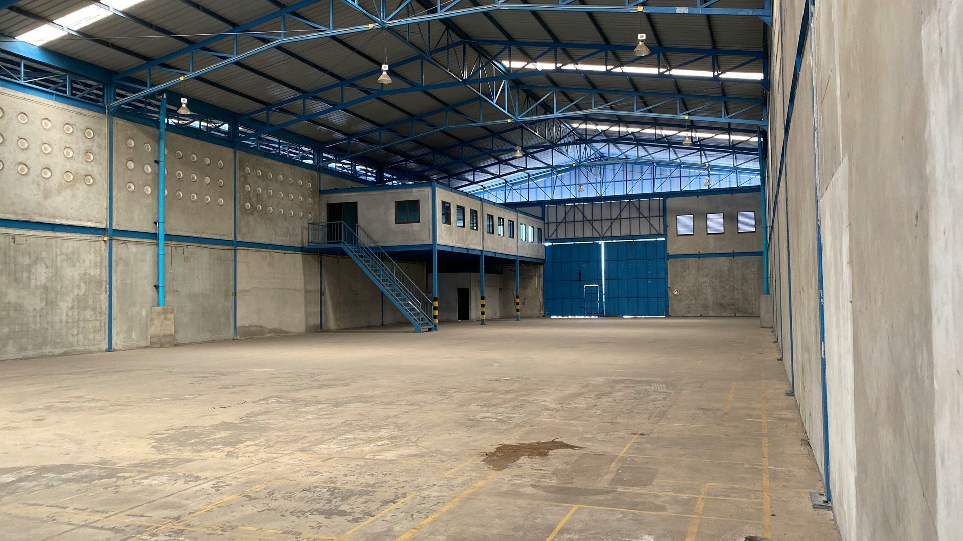 Empty warehouse interior with concrete floor, high ceiling, and elevated office structure. Blue accents.