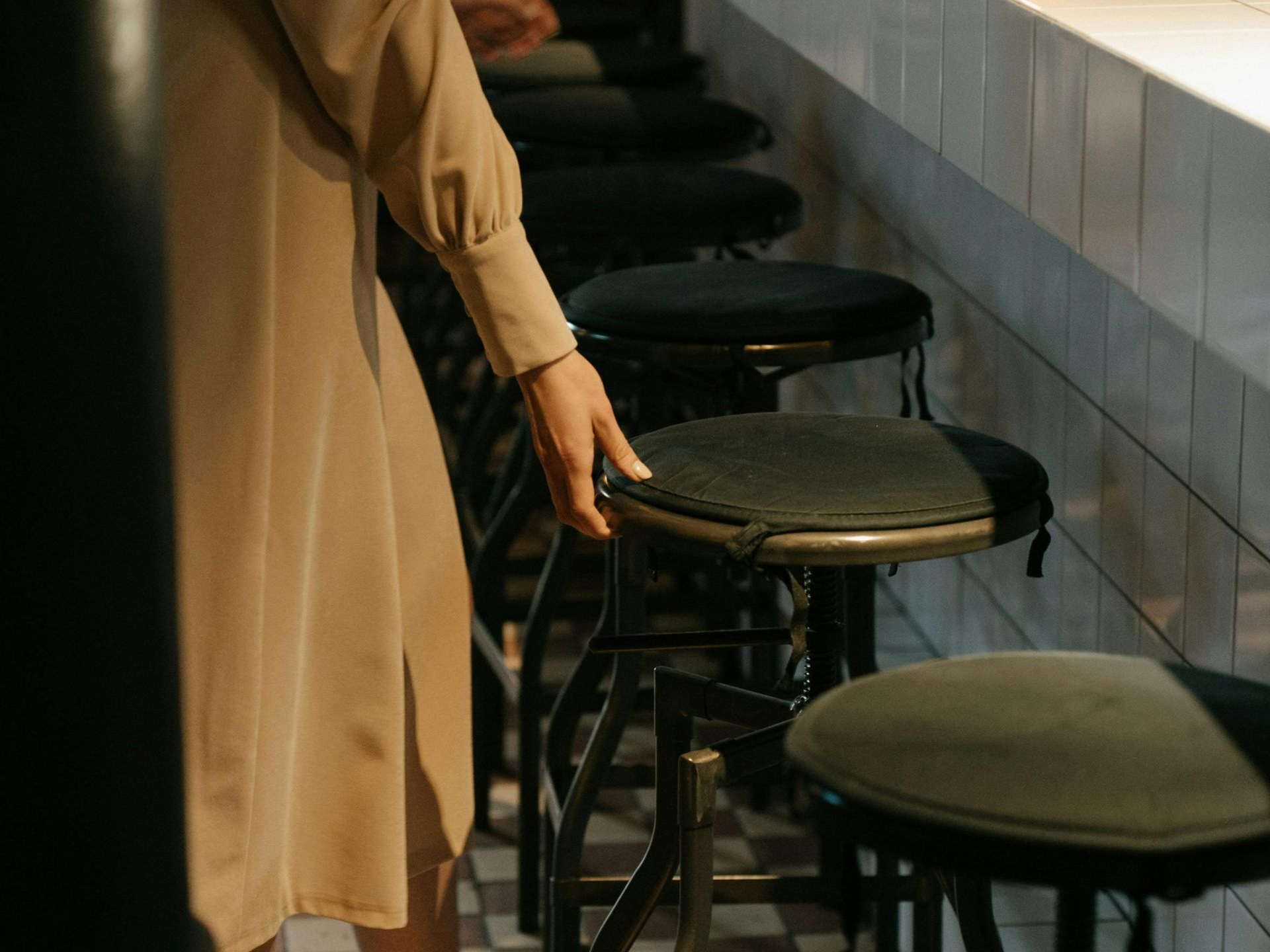 Woman's hand on a black stool in a row; beige dress; sunlight.