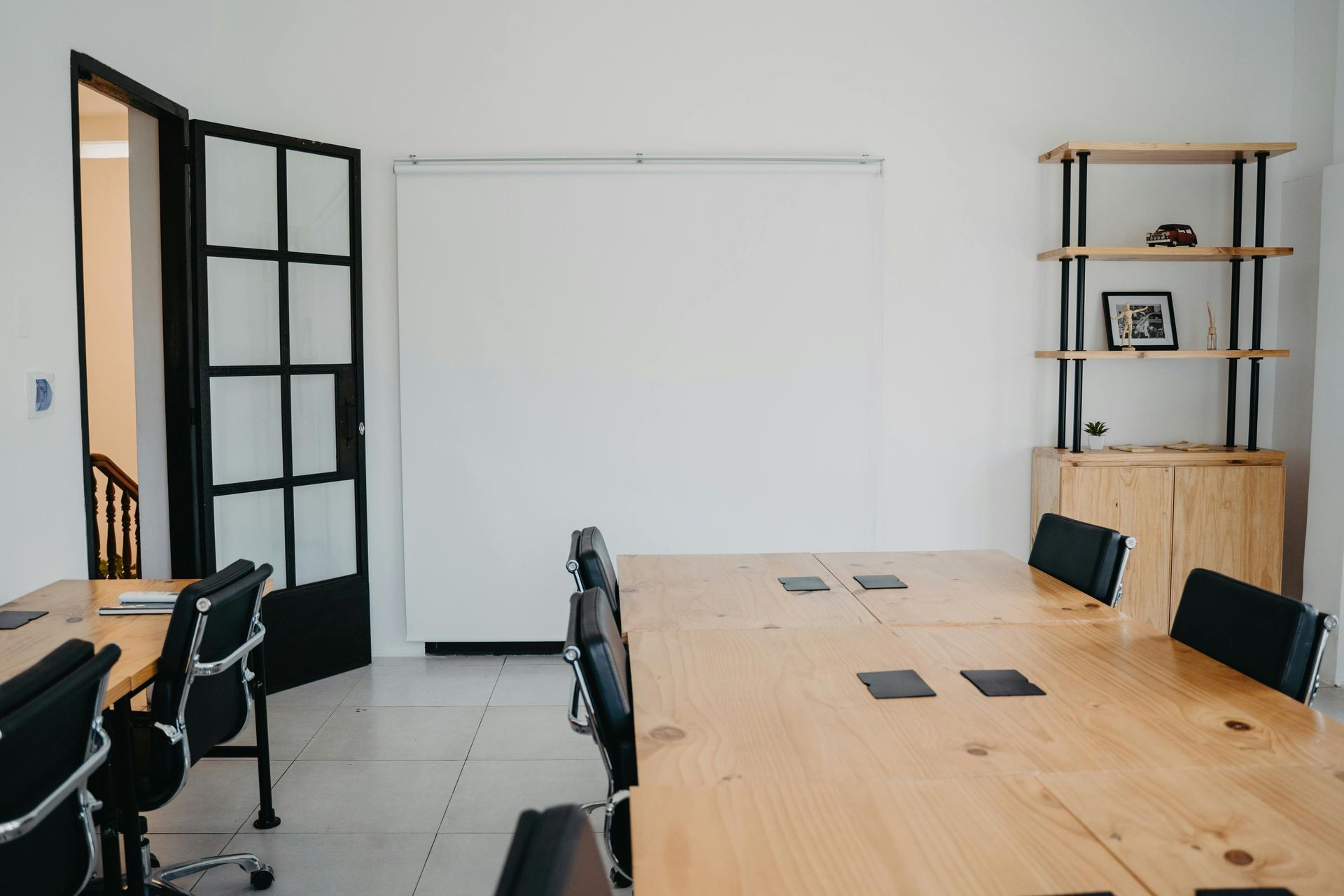 A modern office meeting room with wooden tables, black chairs, a large projection screen, and a wooden display shelf.