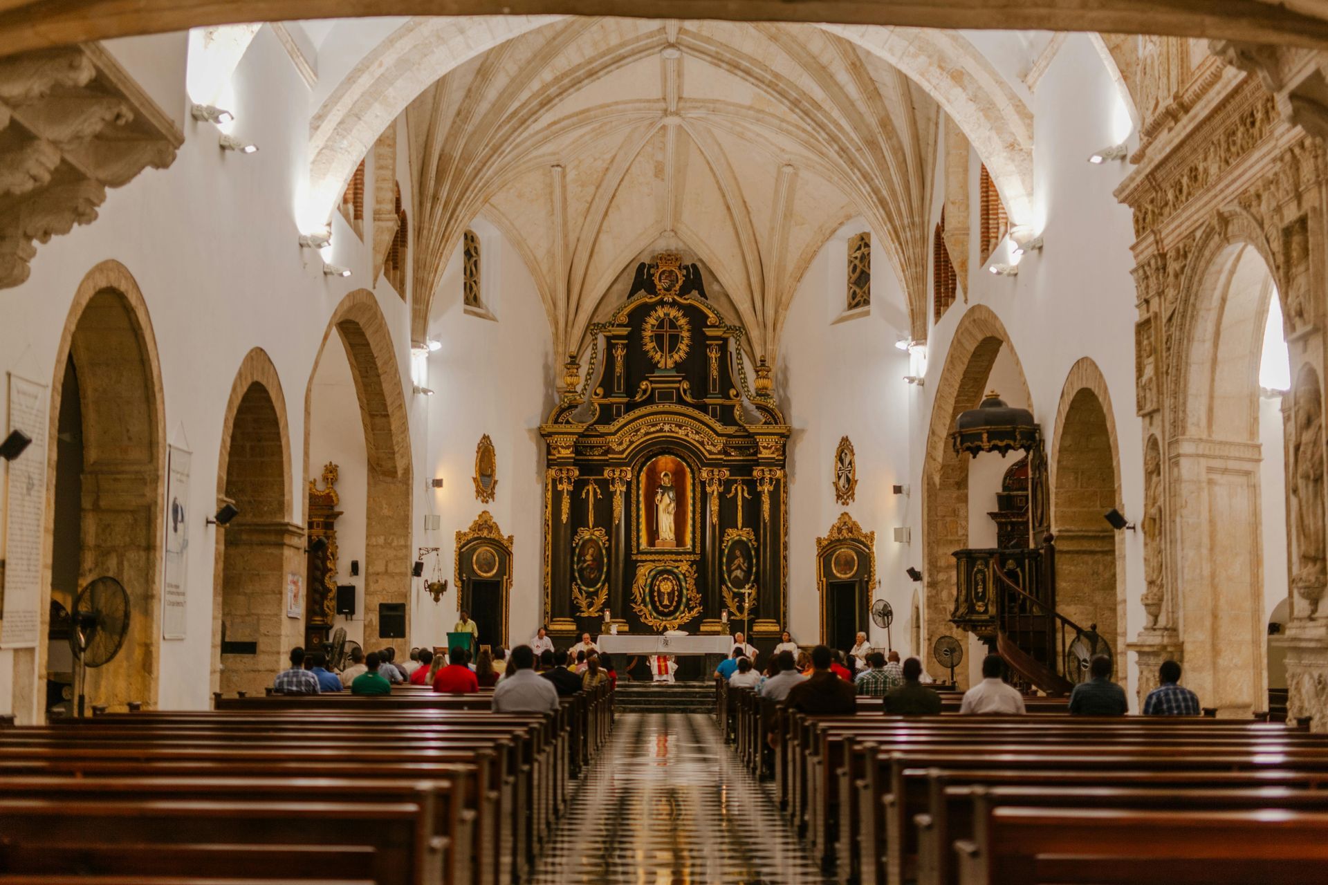 Interior of a church with arched ceilings and ornate golden altar. People sit in pews facing the altar.