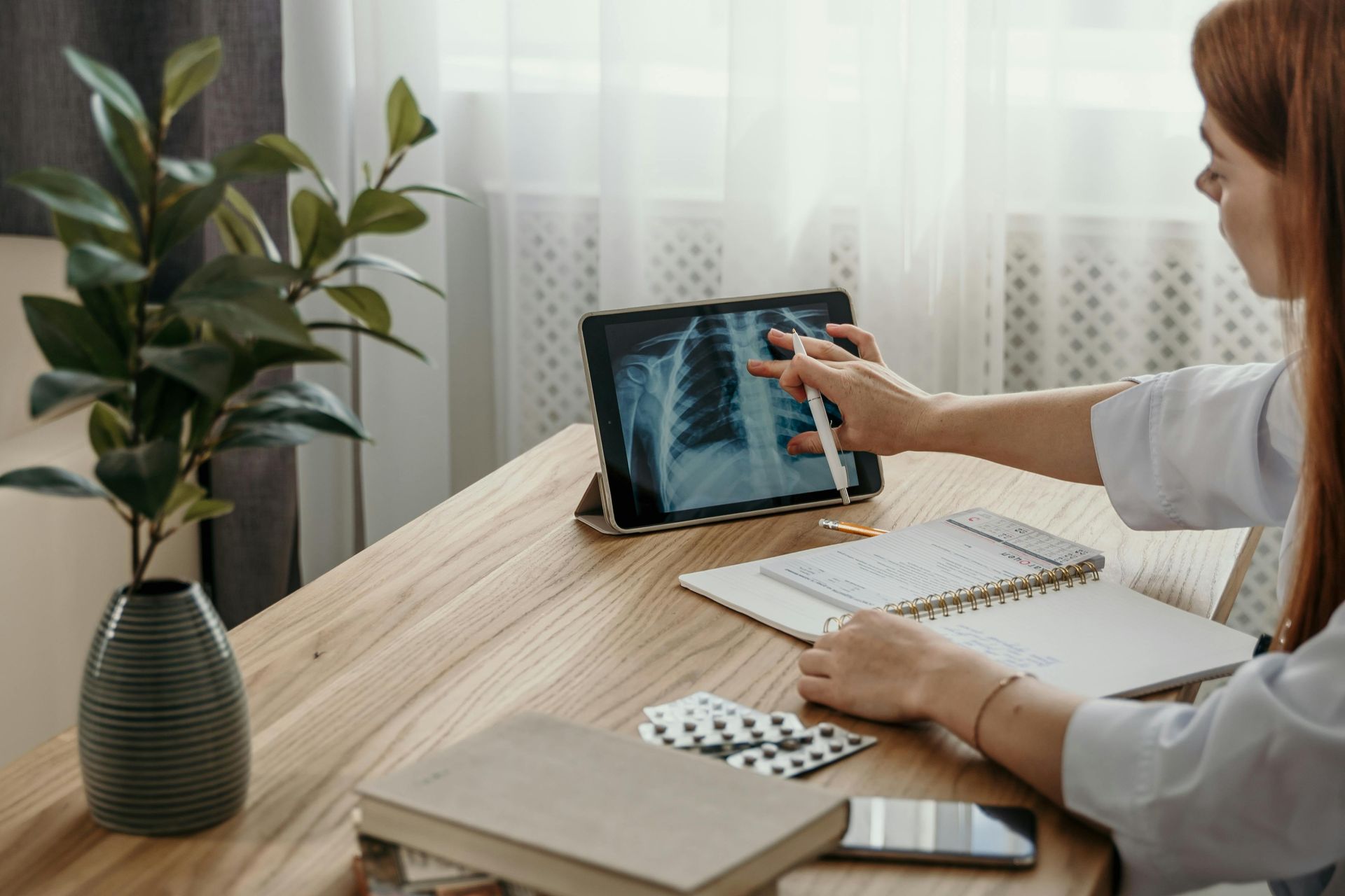 Woman in a white coat examines an X-ray on a tablet, pointing with a stylus. Notepad and medication on desk.