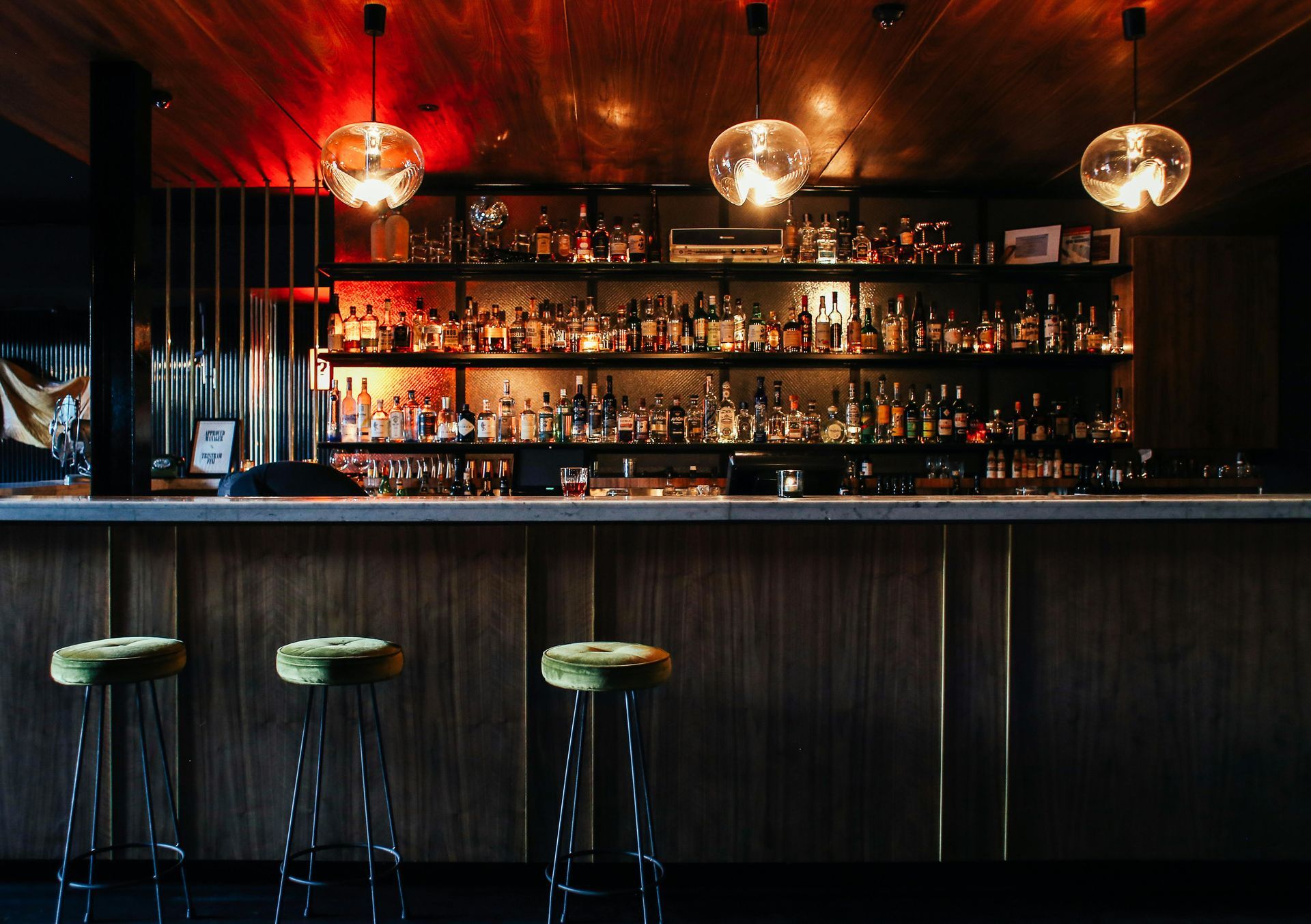 Bar with shelves of liquor, green stools, and round pendant lights.