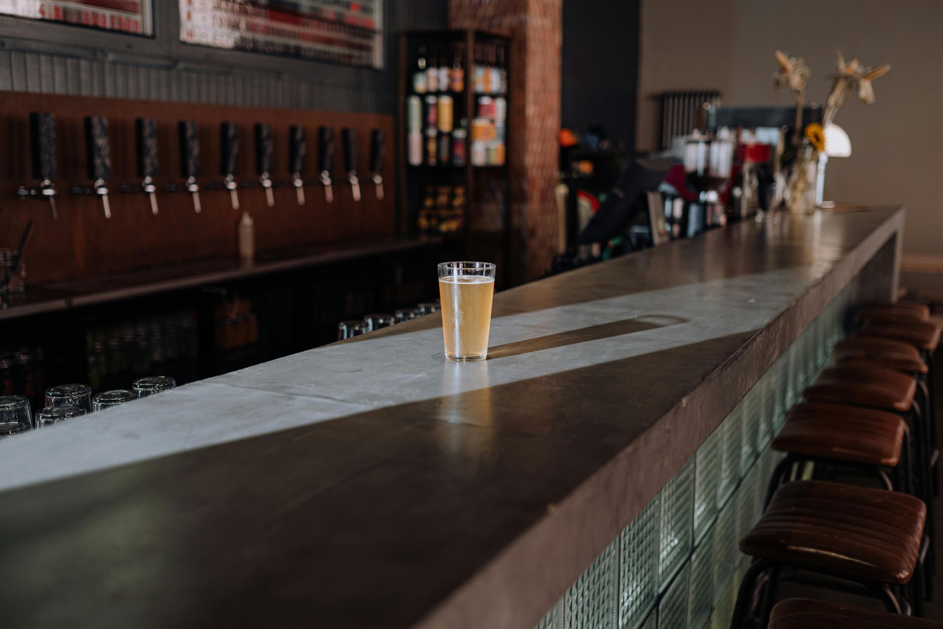 A glass of beer sits on a bar in a dimly lit brewery, with tap handles and stools.