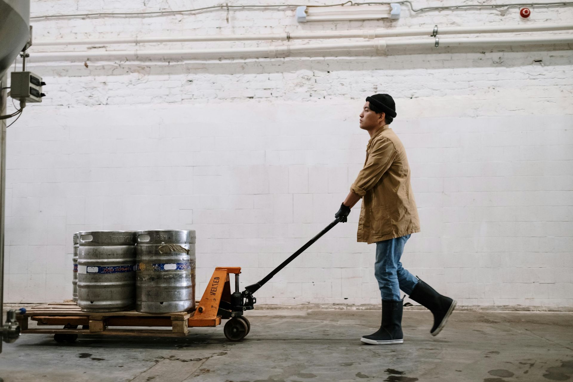 Man in boots pulls a pallet loaded with two kegs using a hand truck in a warehouse.