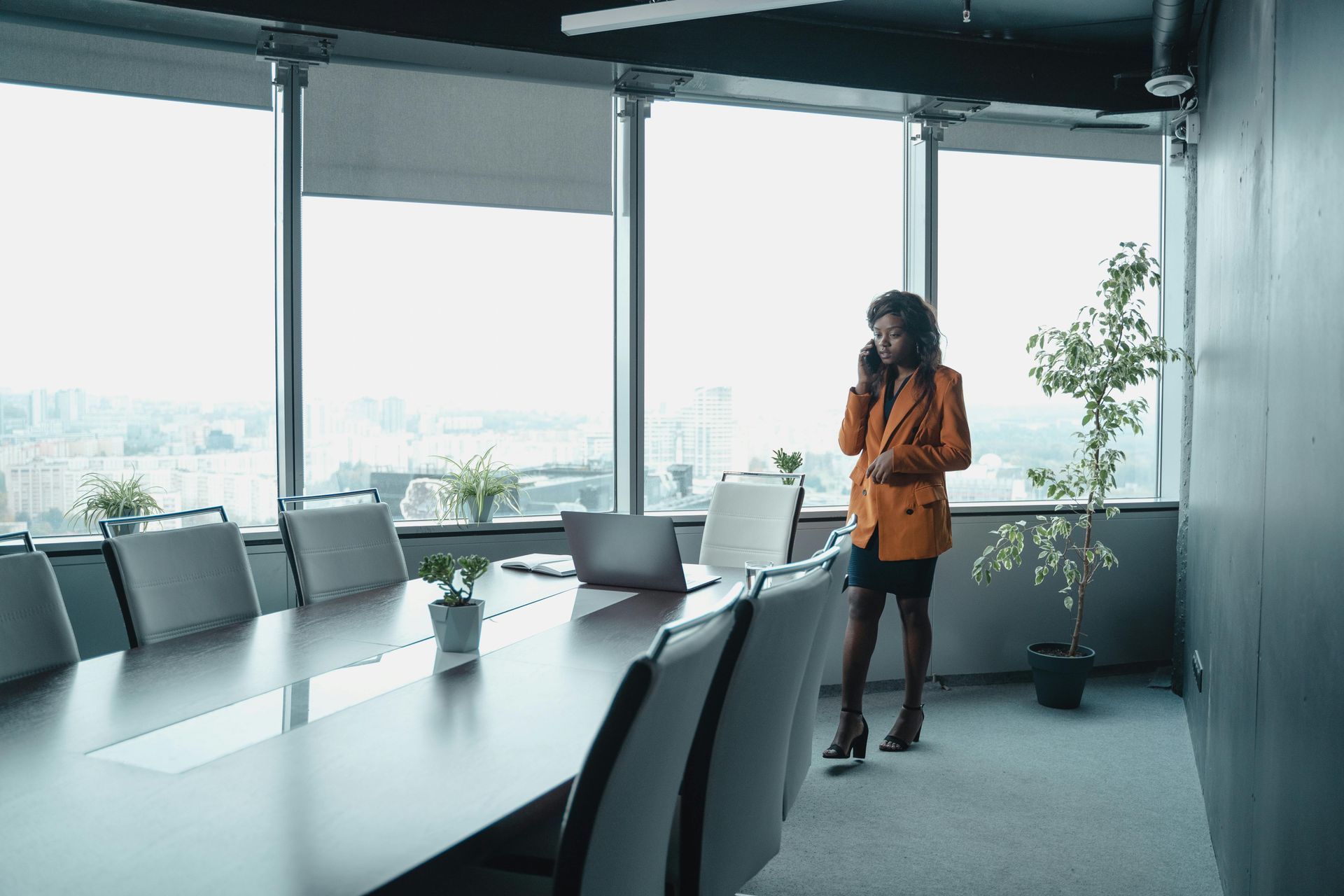 Woman in orange blazer on phone in modern office with a conference table and cityscape view.