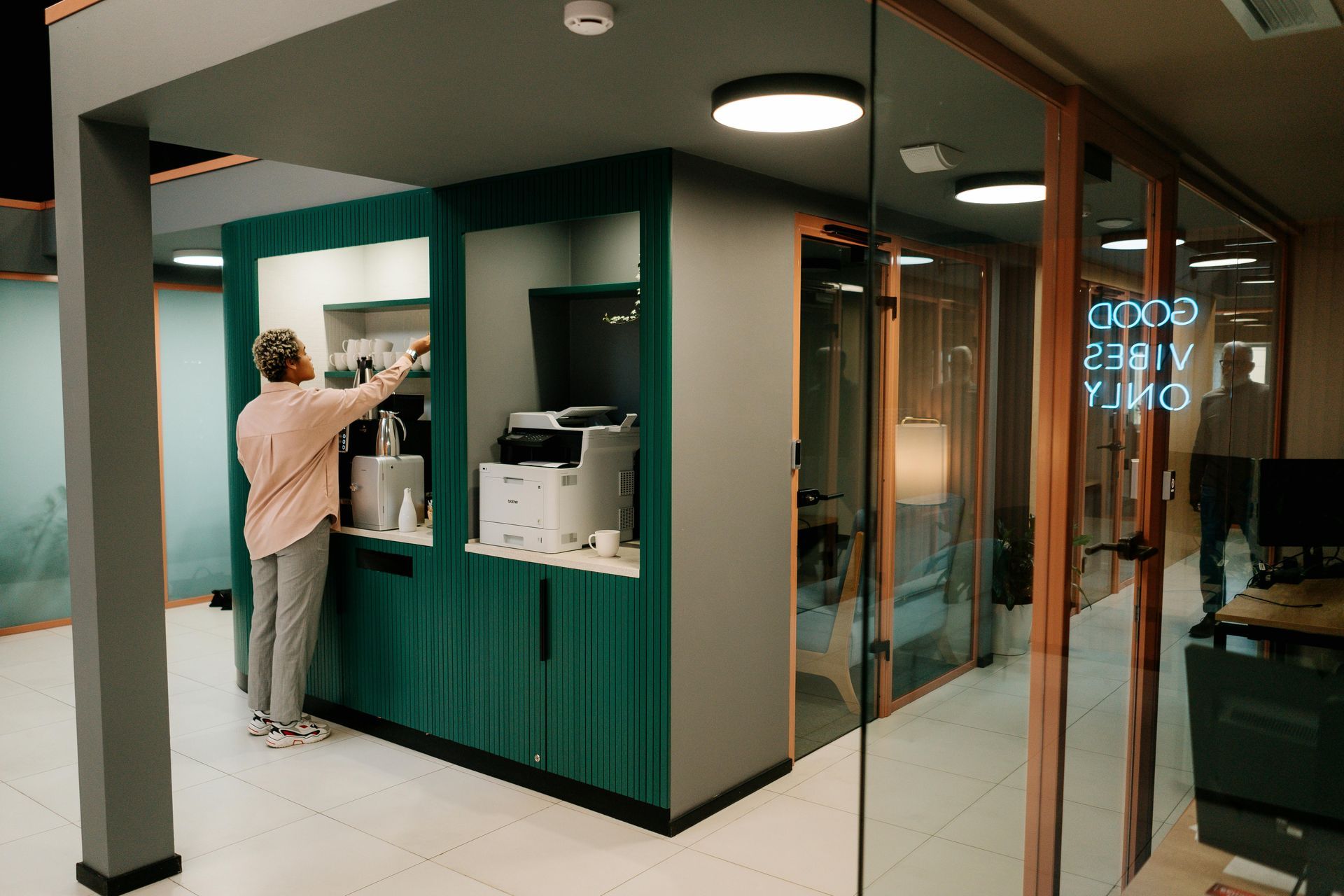 Person at a green and gray office kitchenette, near glass-walled office with 