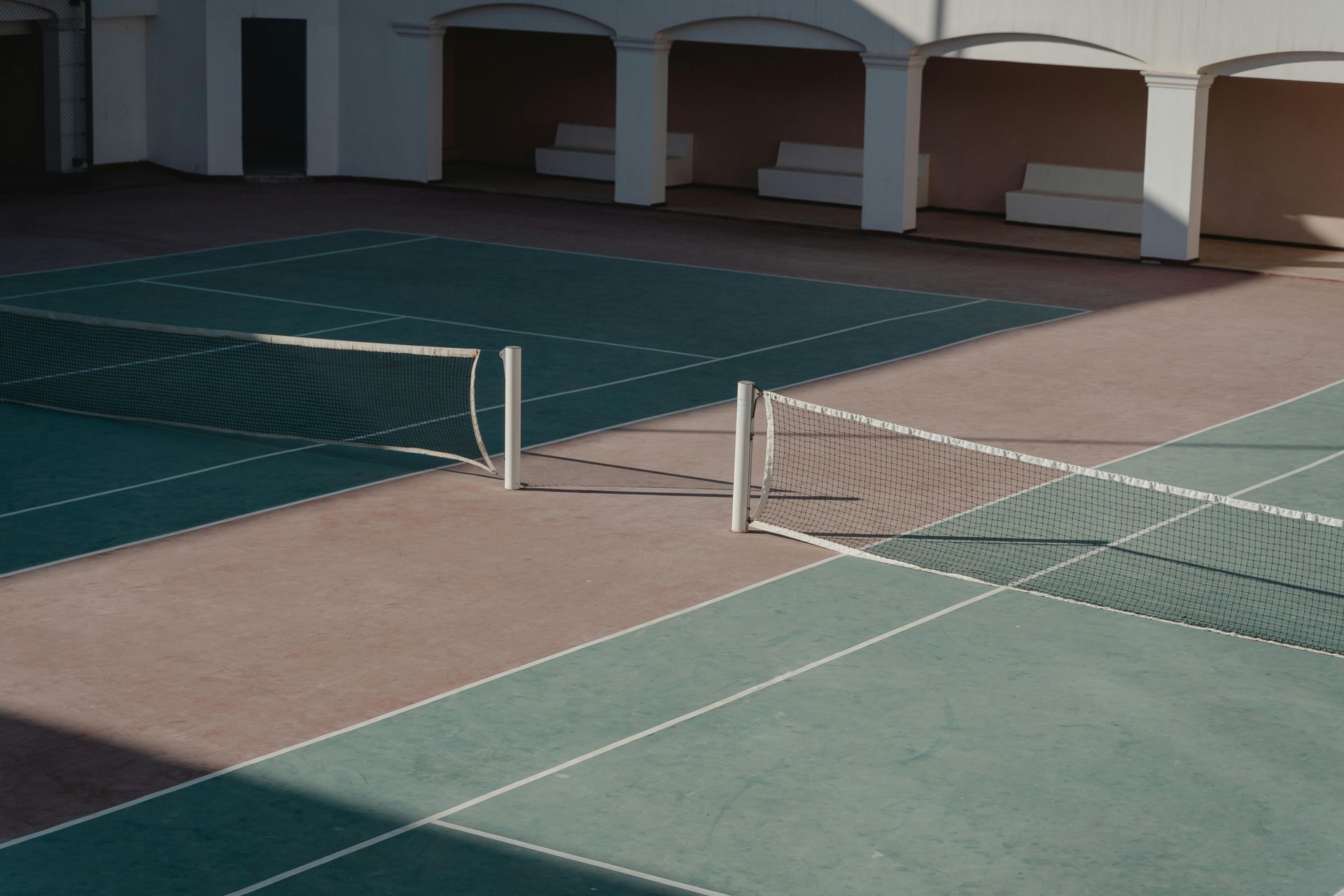 Empty tennis court with net, brown and green court surfaces, white colonnade in background.