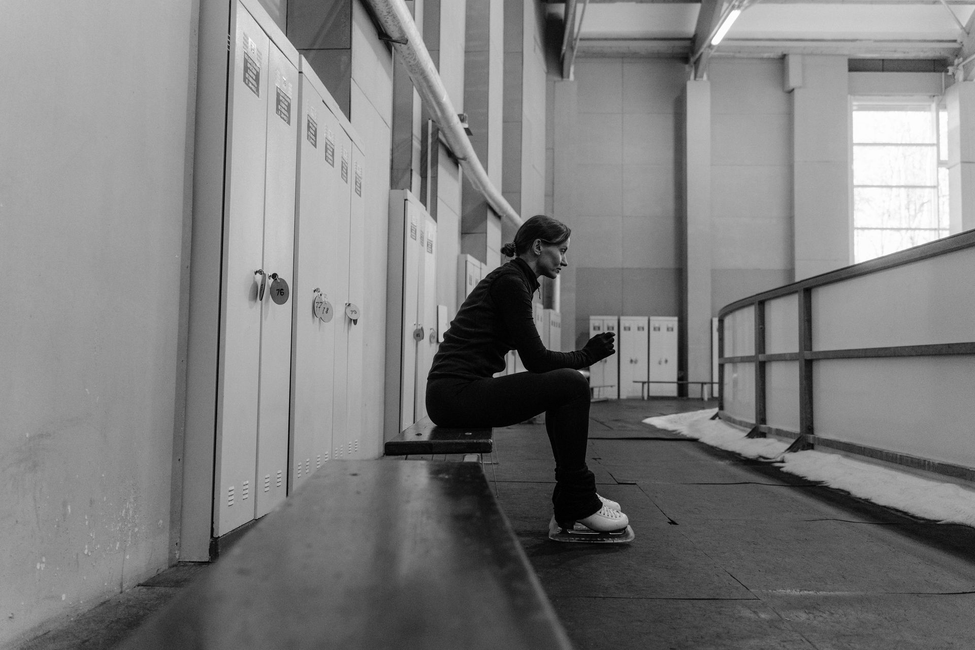 Figure skater seated on a bench, wearing skates, looking at their phone in an ice rink locker room.