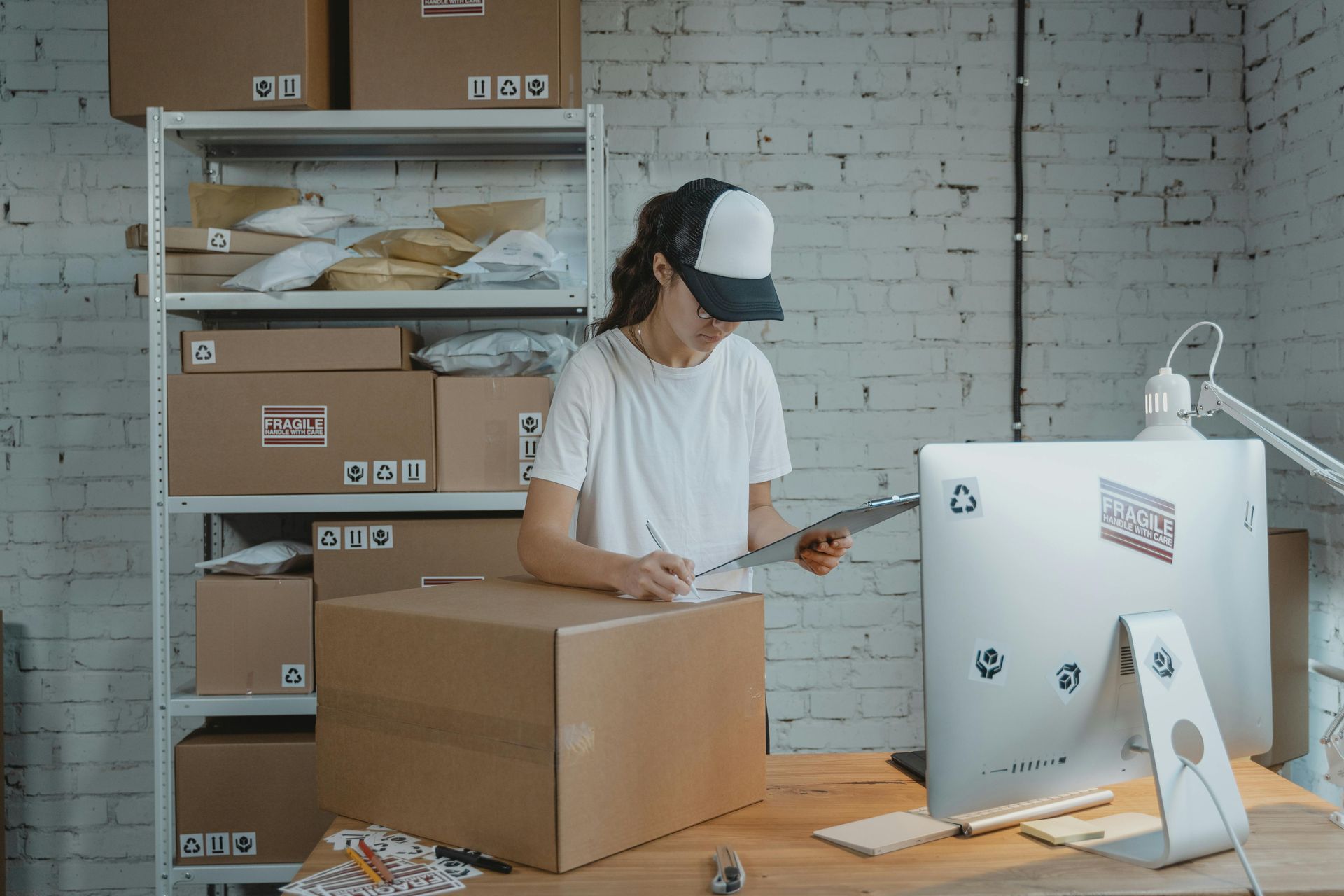 Woman in cap, writing on clipboard next to cardboard box, desk with computer and shelves of packages in a warehouse.