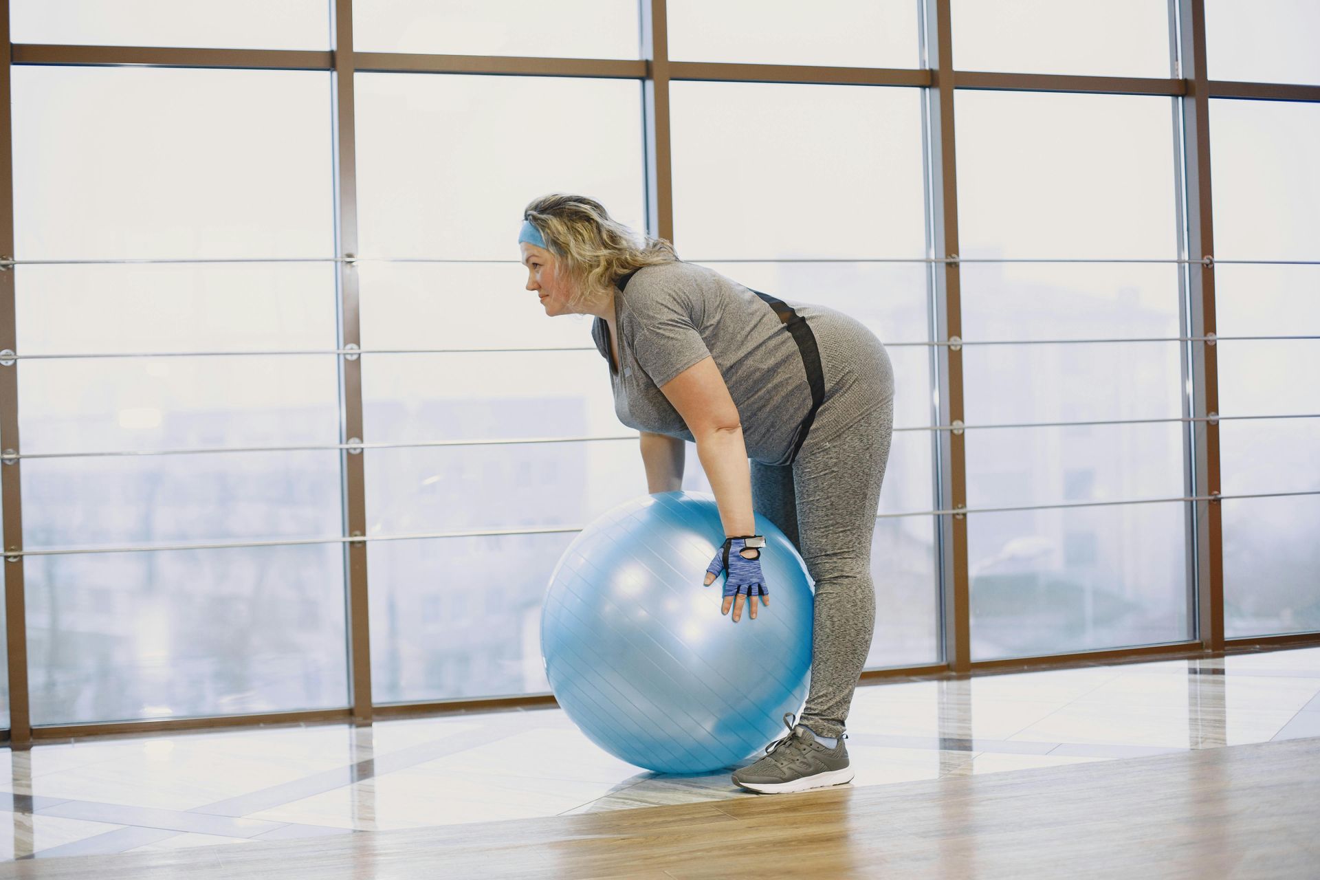 Woman in gray workout clothes leaning over a blue exercise ball in a gym.