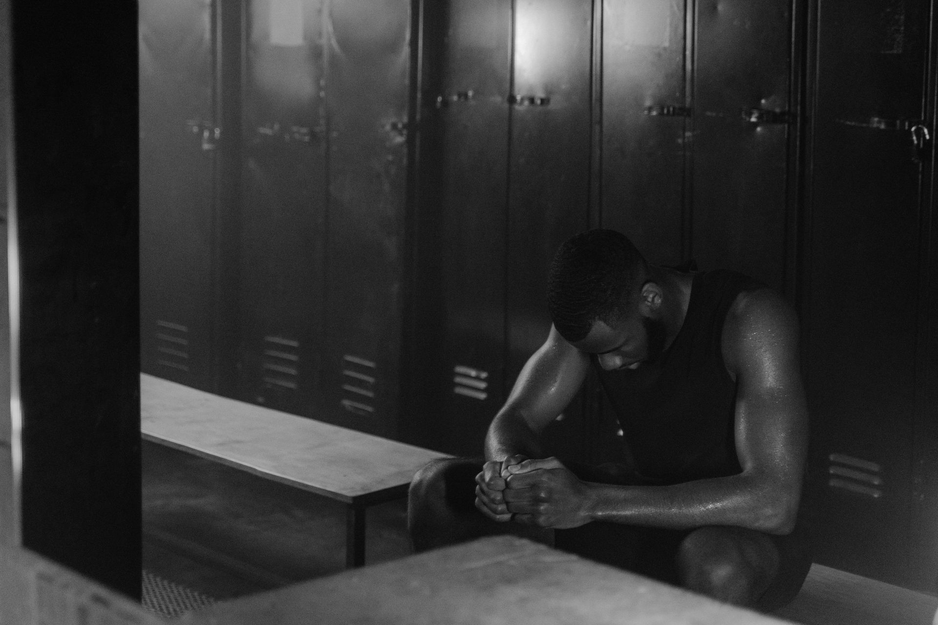 Athlete in a dark locker room, exhausted. Sweat on skin, hands clasped. Sitting on bench with lockers in background.