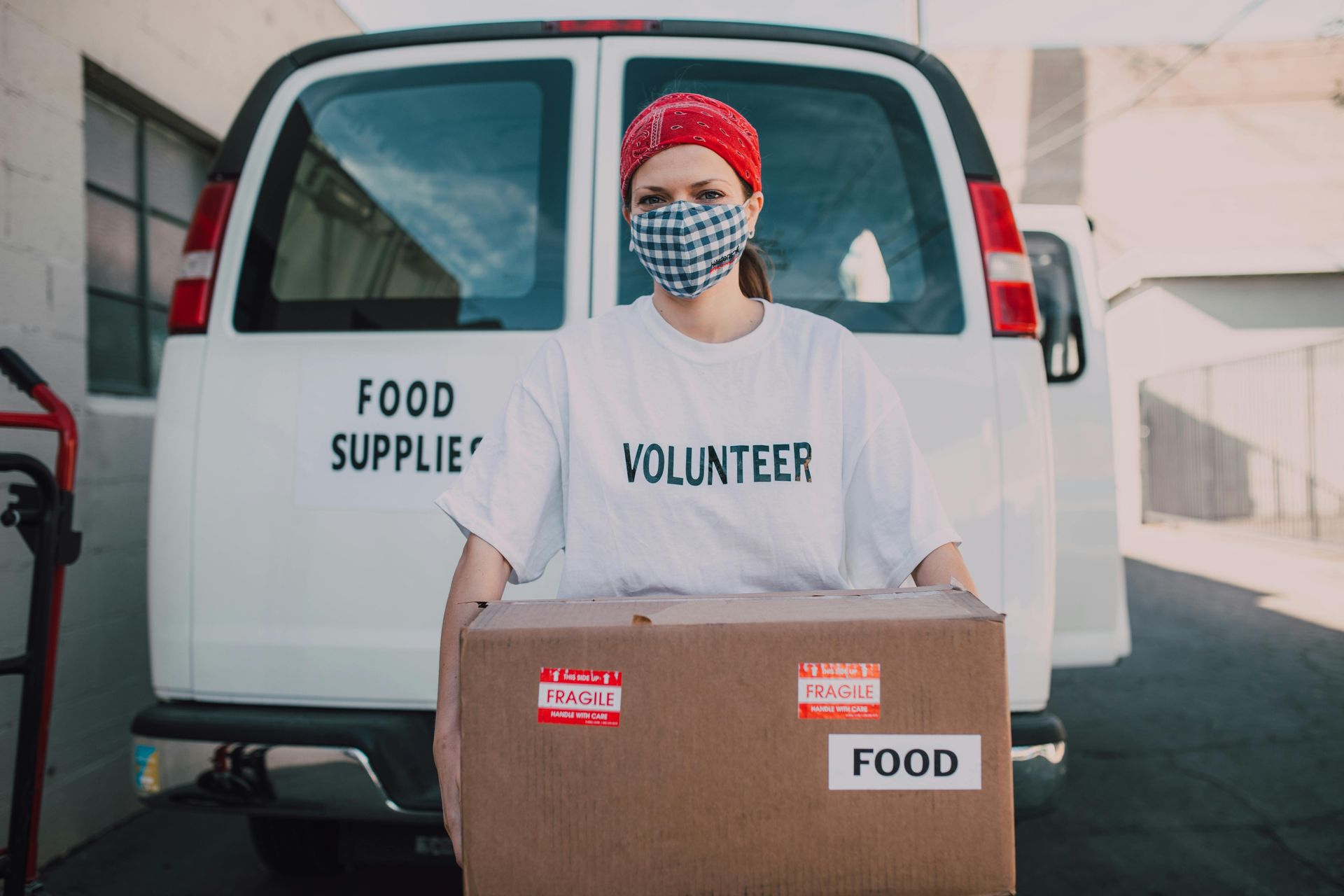 Volunteer in a face mask carrying a box of food in front of a van labeled