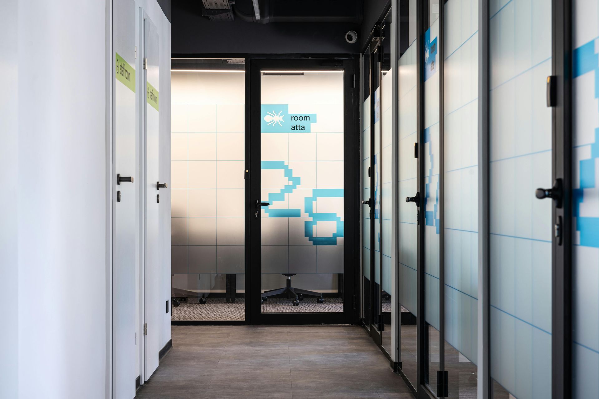 Narrow office hallway with white doors and frosted glass panels; a room with blue pixelated design is at the end.