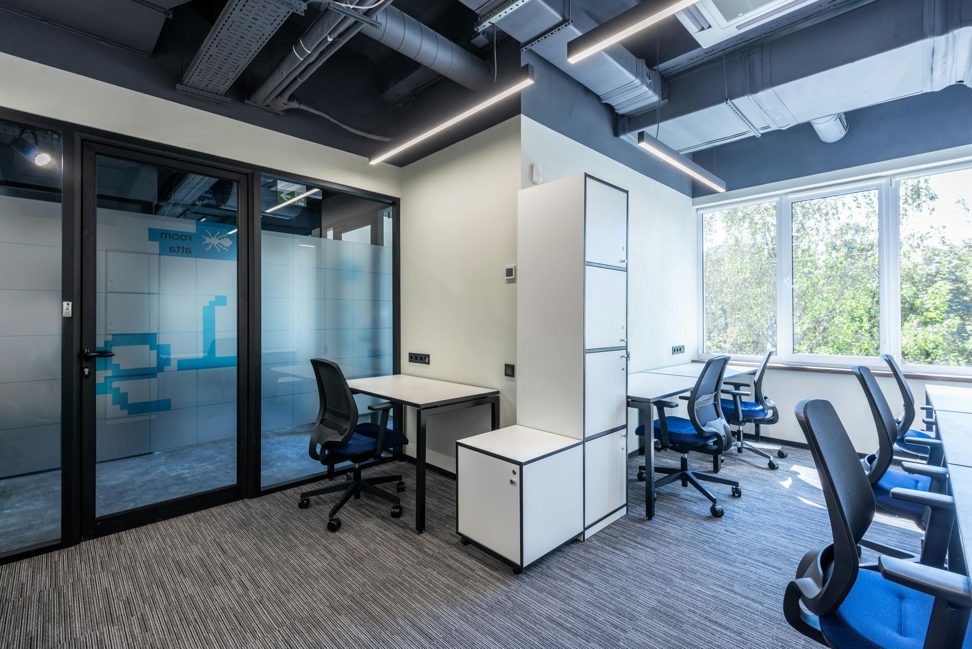 Modern office interior with desks, chairs, and glass-walled rooms. Natural light from large windows.