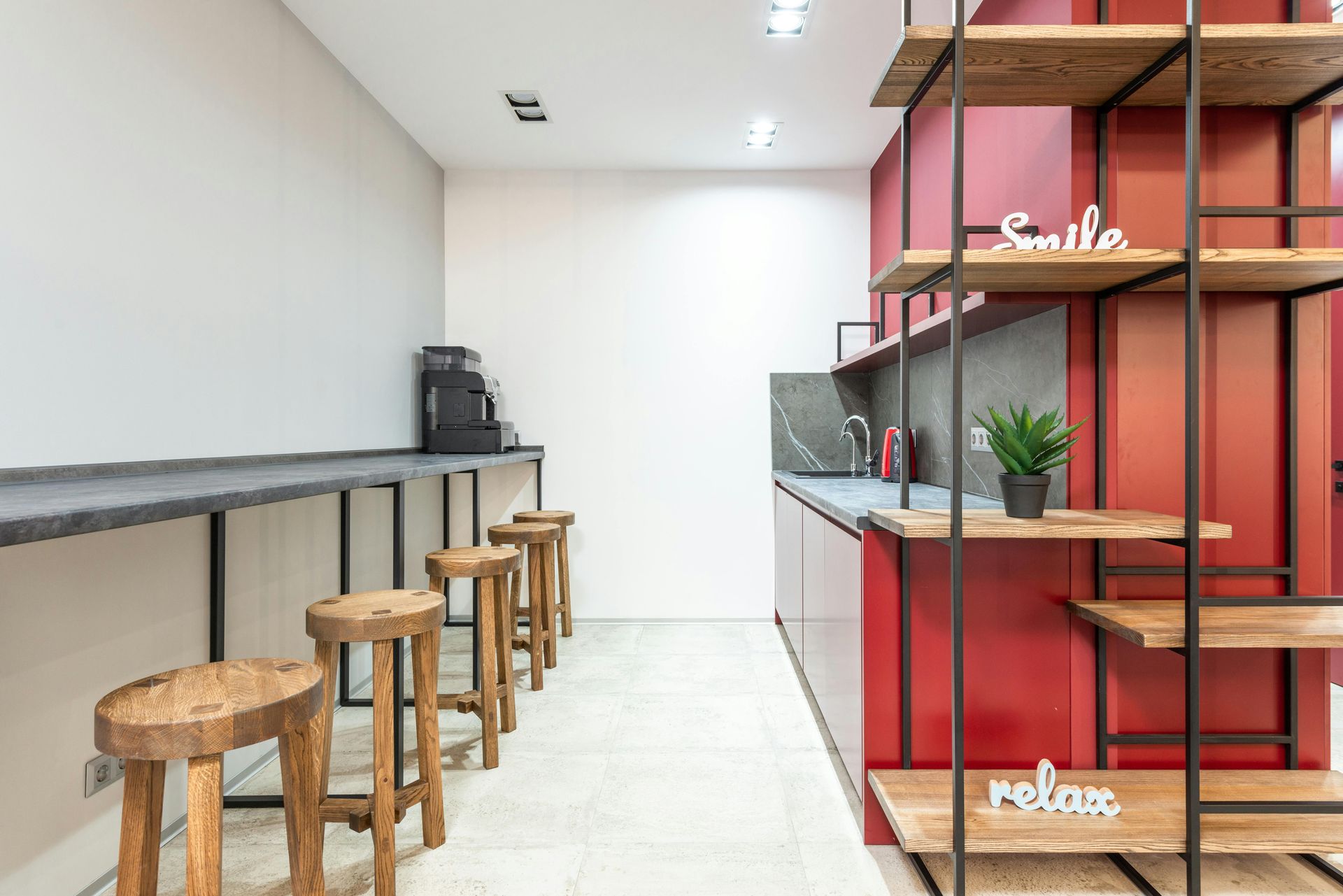 Modern office kitchen with stools, espresso machine, red shelving, and minimalist decor.