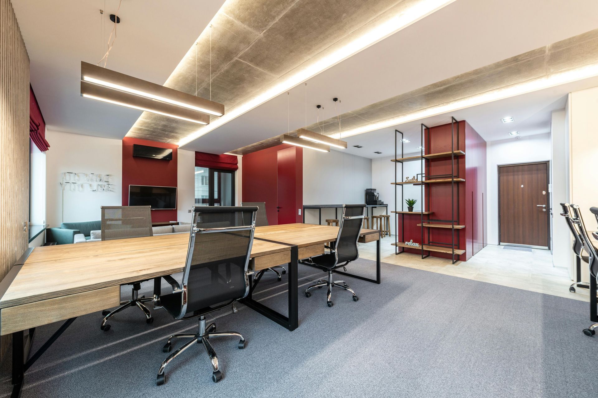 Modern office space with desks, chairs, and red accents. Features concrete ceiling, wood elements, and grey carpet.